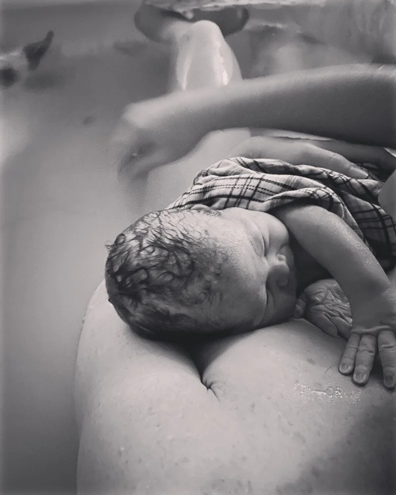 A newborn baby with wet hair is cradled on an adult's shoulder in a black-and-white photo, with the adult's arm visible.