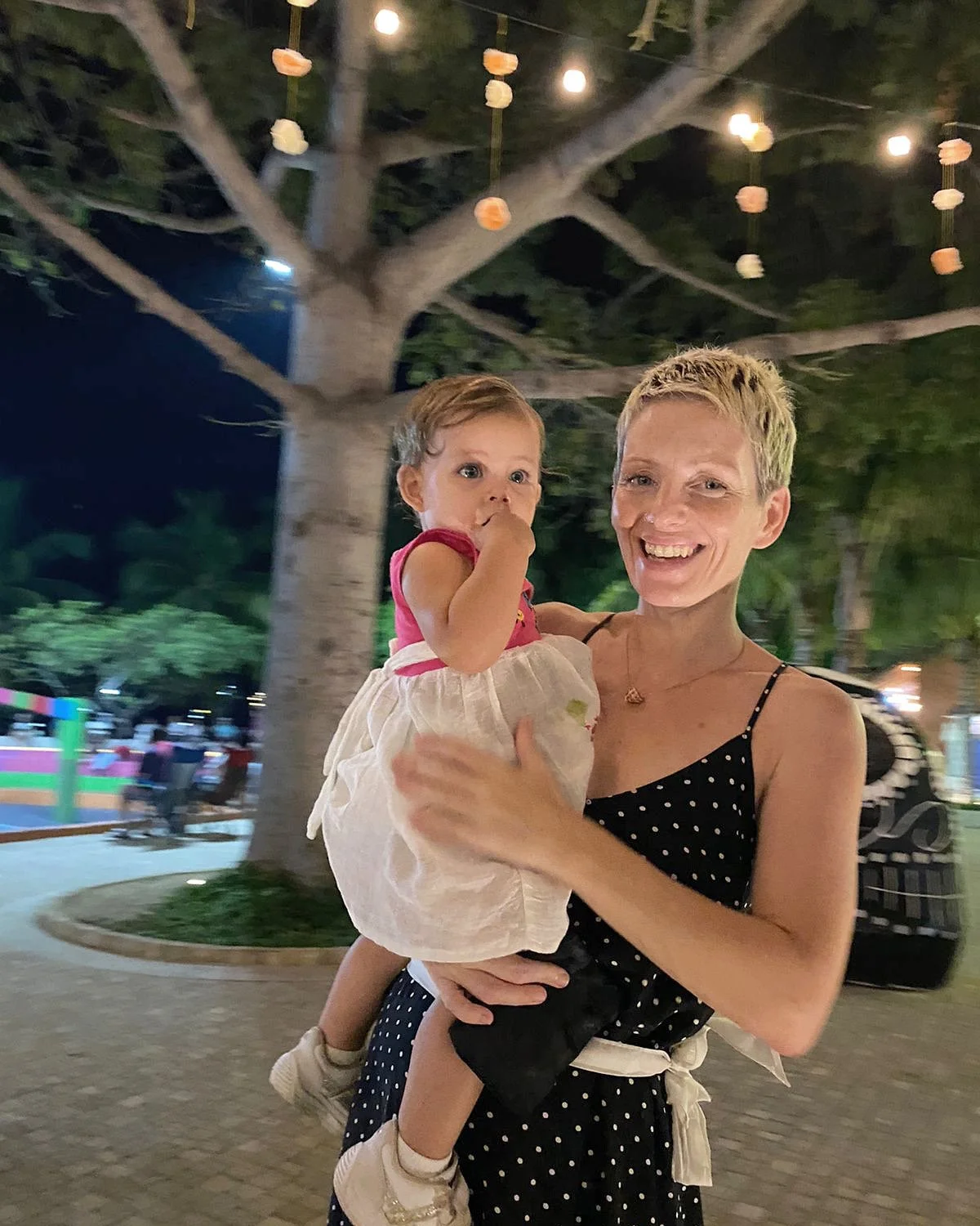 A woman holding a young girl with a background of a large tree decorated with hanging lanterns, nighttime setting, and colorful lights in the distance.