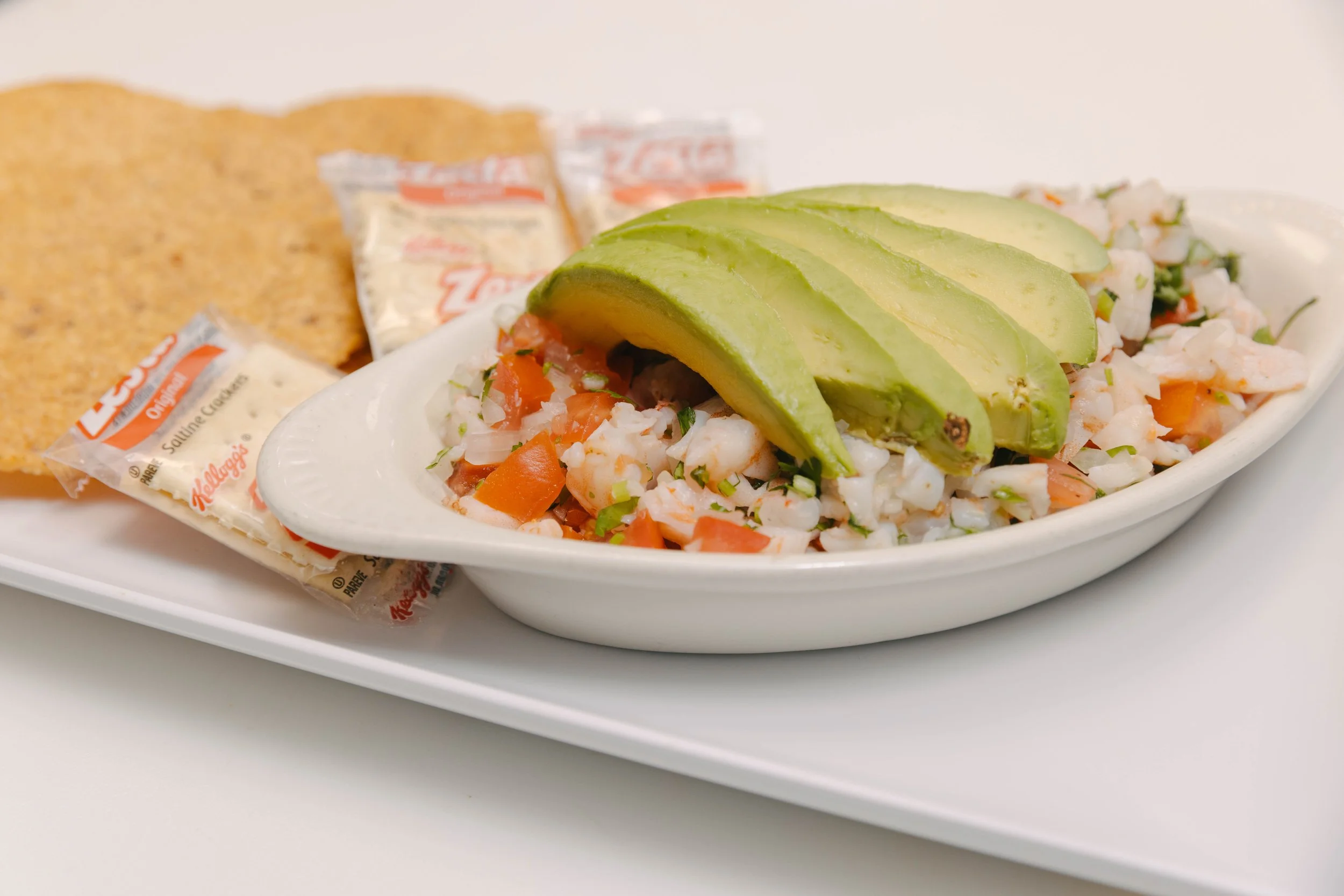 A bowl of seafood ceviche topped with sliced avocado, served with tortilla chips and small packets of saltine crackers in the background.