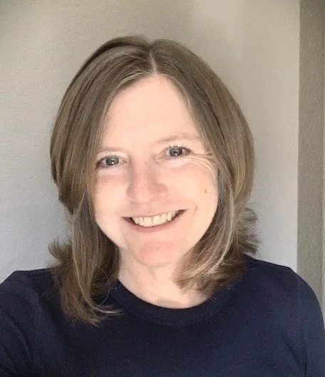 Smiling person with short hair wearing a dark shirt against a light wall.