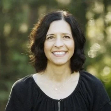 Woman smiling outdoors with blurred greenery in the background.