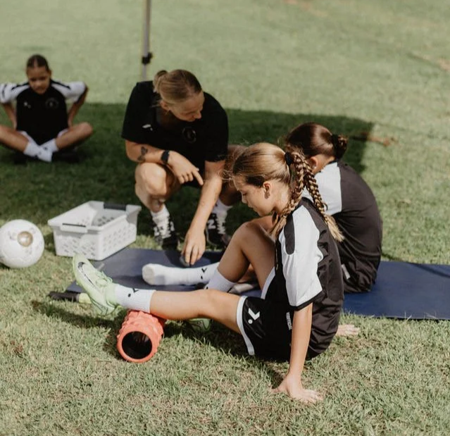 Youth soccer players stretching on a grassy field with a coach, soccer ball, equipment basket, and foam roller