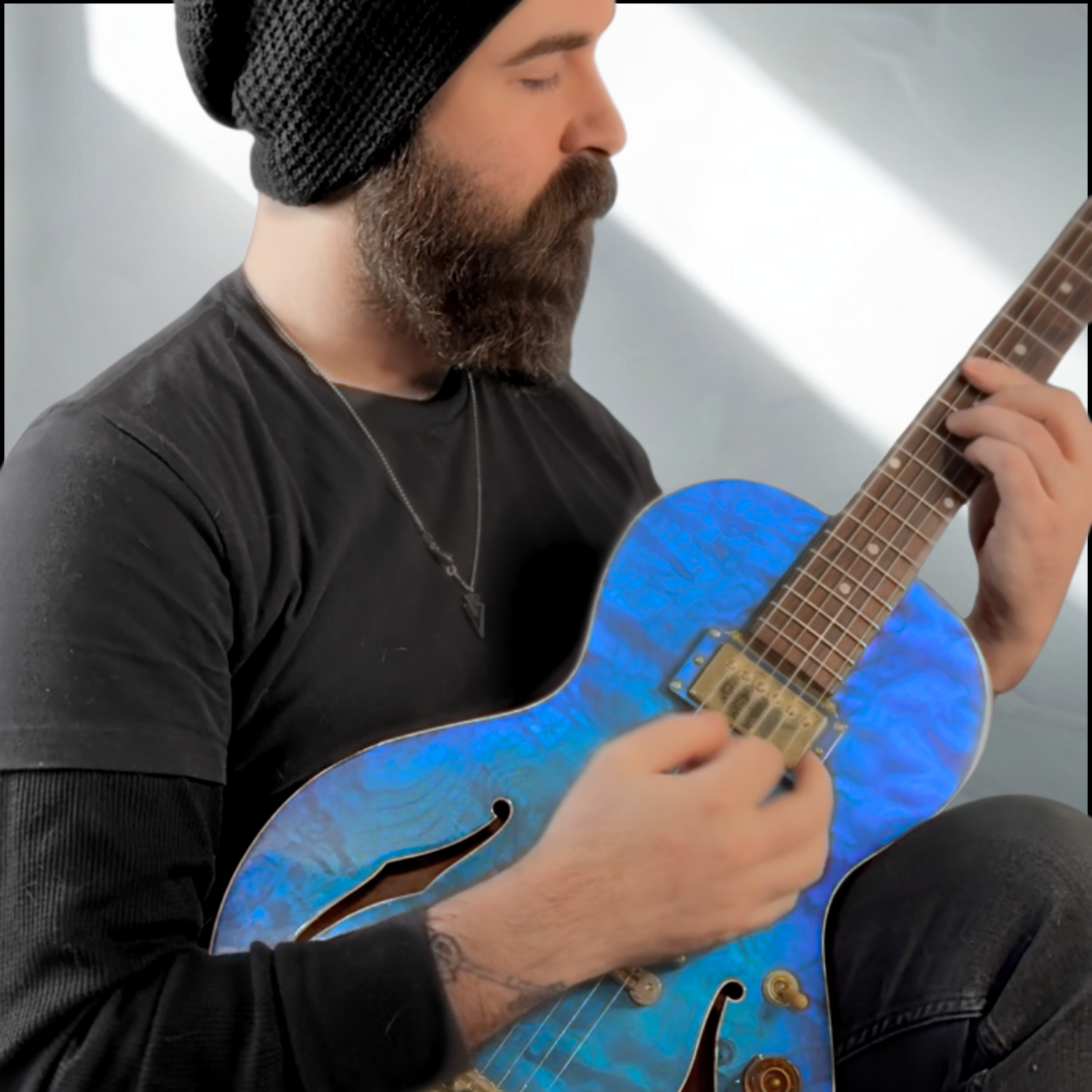 Man with a beard and tattoo playing an electric blue guitar, wearing a black beanie and black shirt.