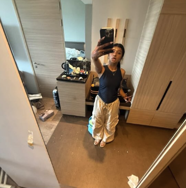 A young woman taking a mirror selfie in a hotel room or bedroom, dressed in a navy top and beige pants, with furniture and personal items visible in the background.