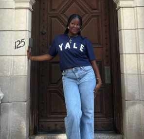 Young woman standing in front of a large wooden door, wearing a navy Yale sweatshirt and blue jeans, smiling at the camera.