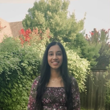 Young woman with long dark hair smiling outdoors in front of green bushes and trees.
