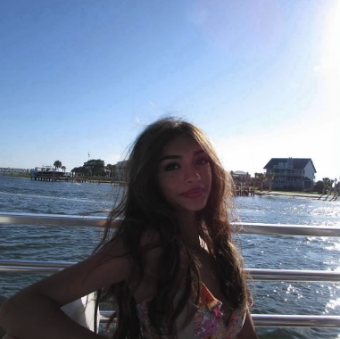 A woman posing by a body of water on a sunny day, with houses and trees in the background.