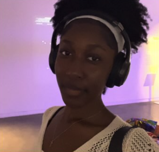 A young woman with natural curly hair wearing headphones takes a selfie indoors with a colorful background.