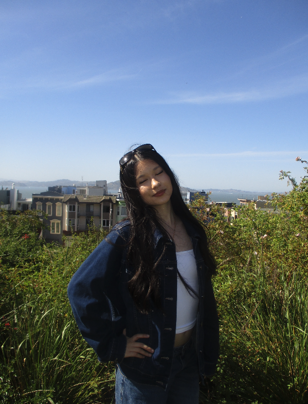 A young woman with long dark hair, wearing a white top and gold jewelry, smiling and standing outdoors with green foliage in the background.