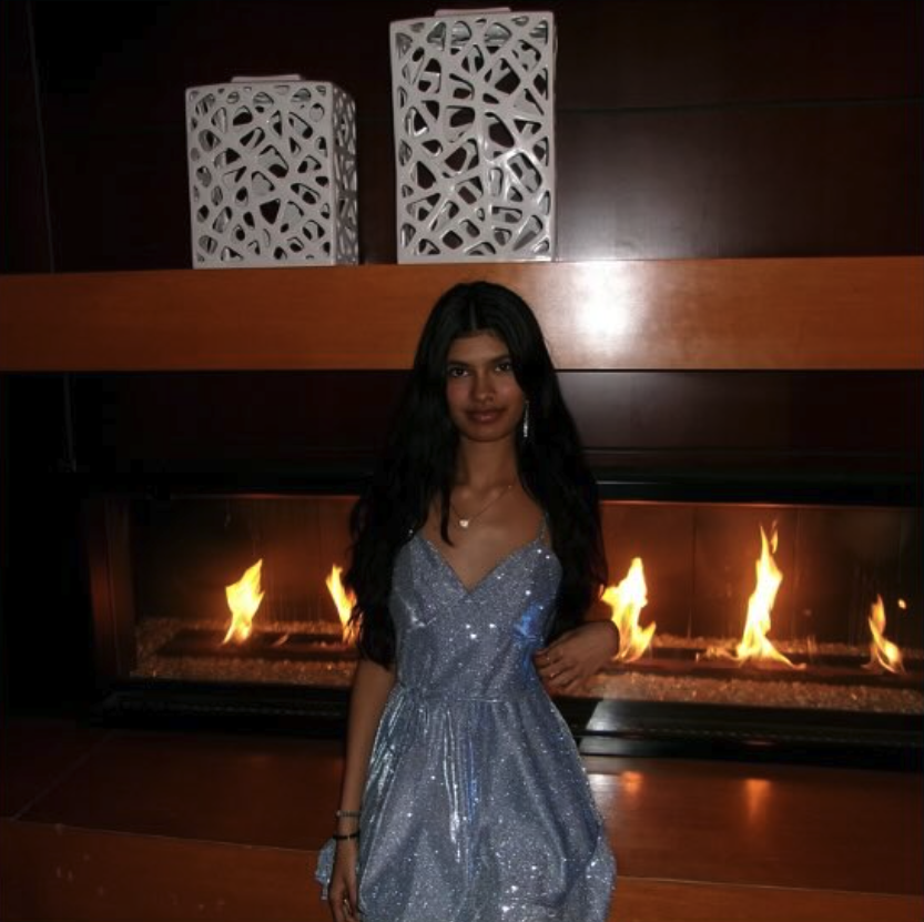 A young woman with long dark hair in a sparkly blue dress standing in front of a modern fireplace with flames and decorative white sculptures above.