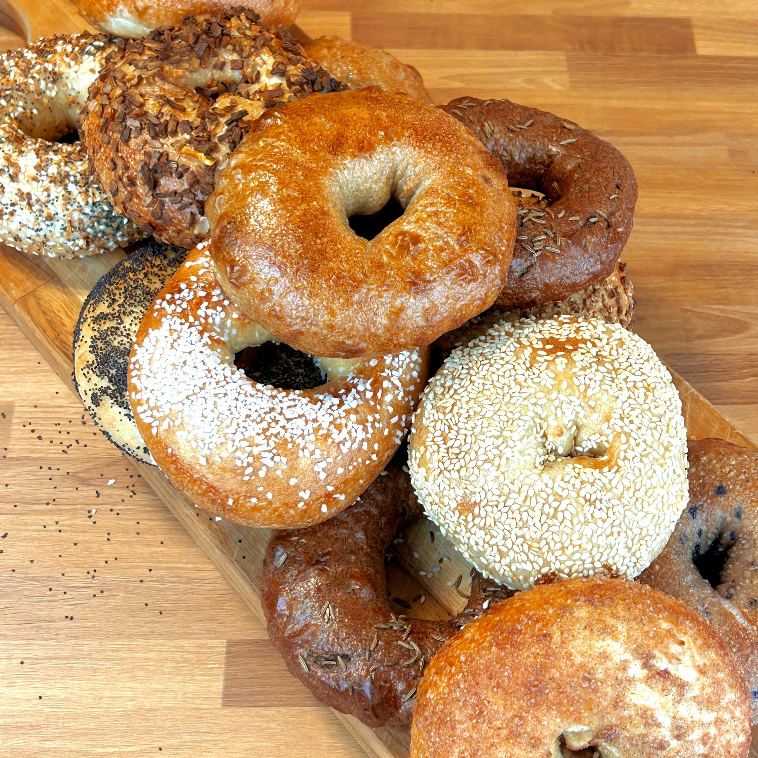 There are 13 bagels sitting on a serving tray on a table. The bagels have various toppings such as sesame seeds, salt, cinnamon sugar, garlic, and onion