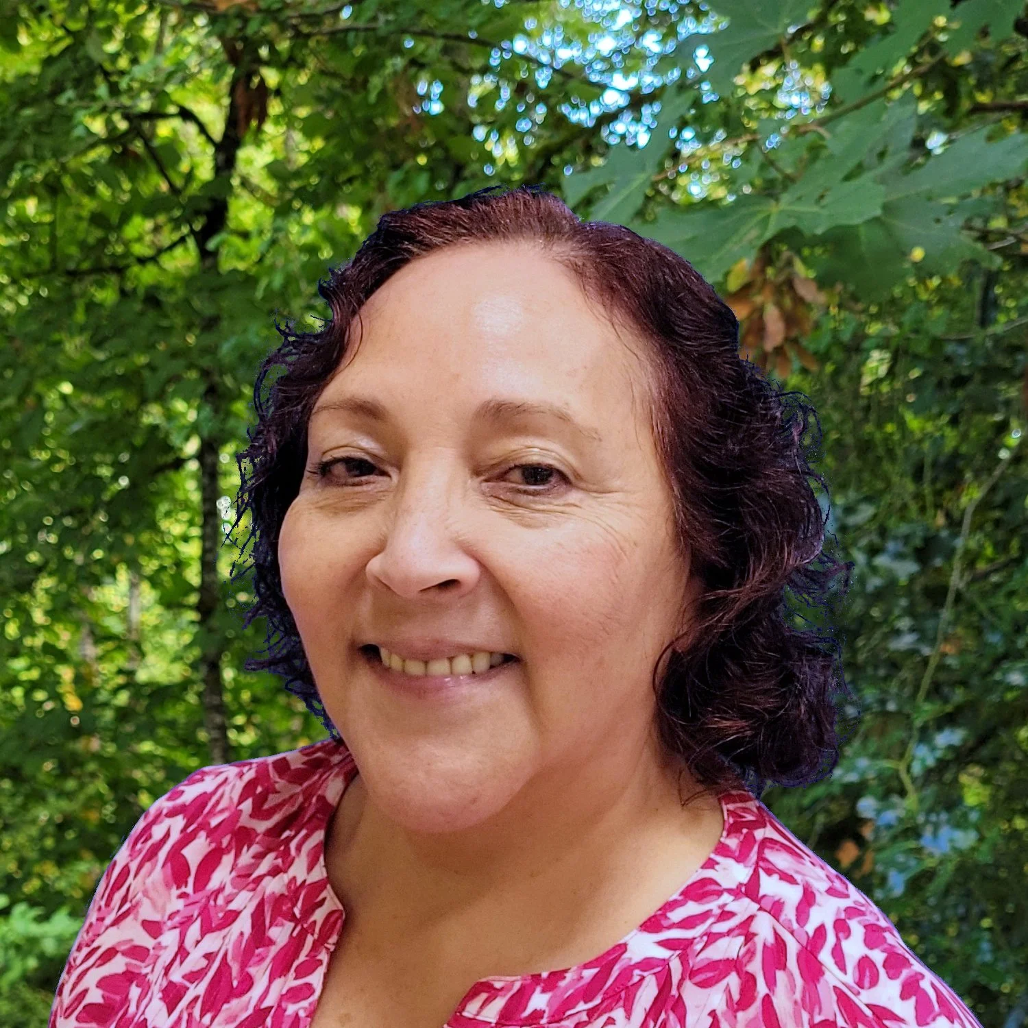 A woman with curly, dark hair and a swirly magent and white shirt smiles in front of a lush green forest