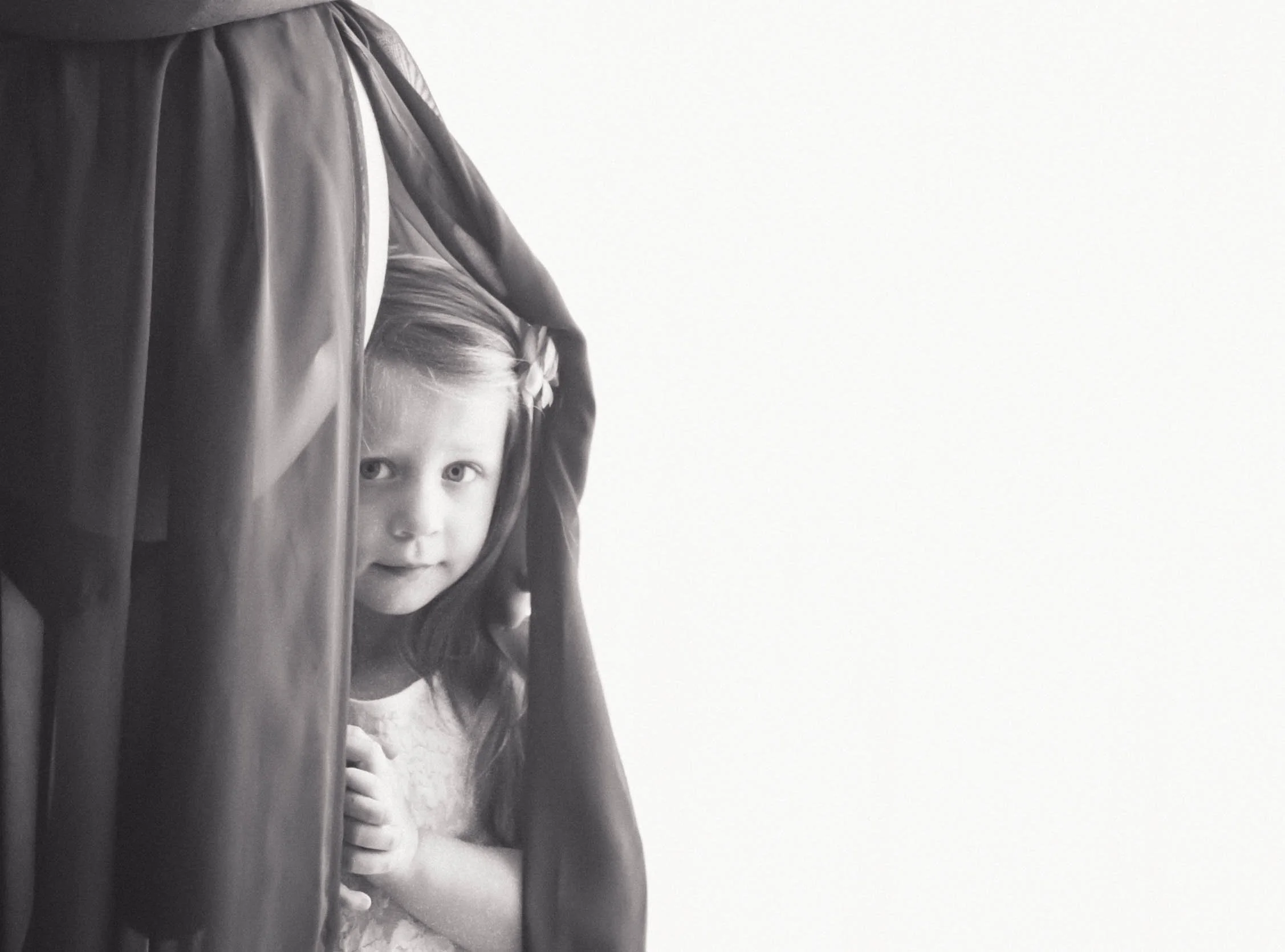 Black and white photo of a young girl hiding behind a person's dress.
