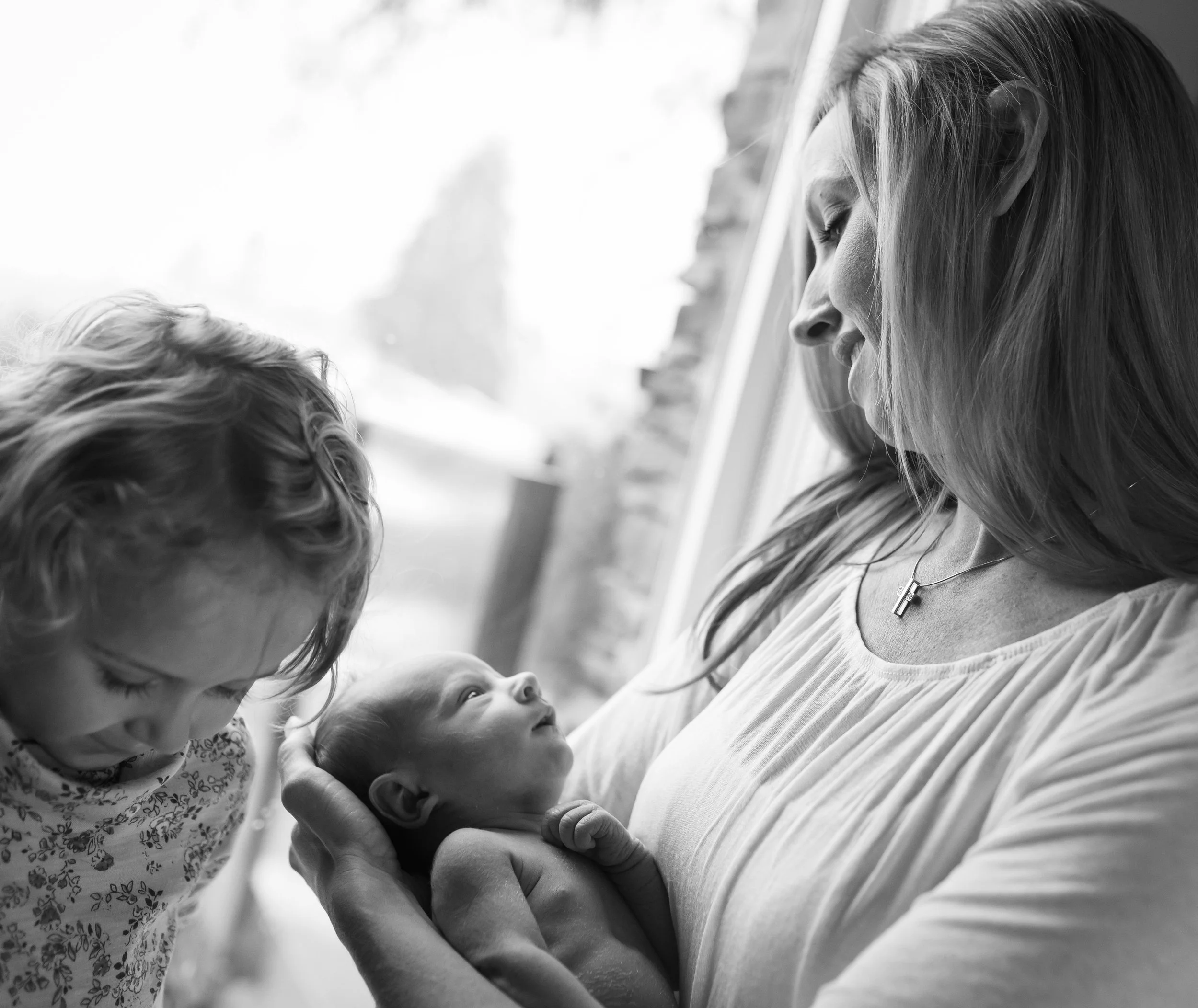 Black and white photo of a woman holding a baby, with a child standing nearby looking at the baby.