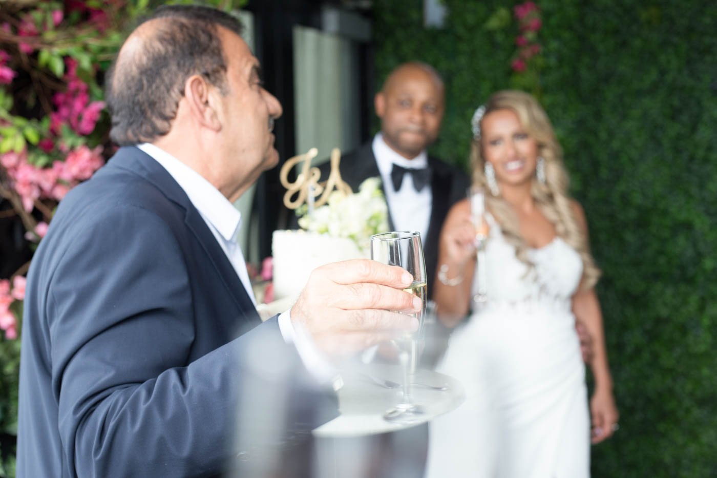 Wedding guest giving a toast with champagne in foreground, while bride and groom smile in background, surrounded by greenery and flowers.