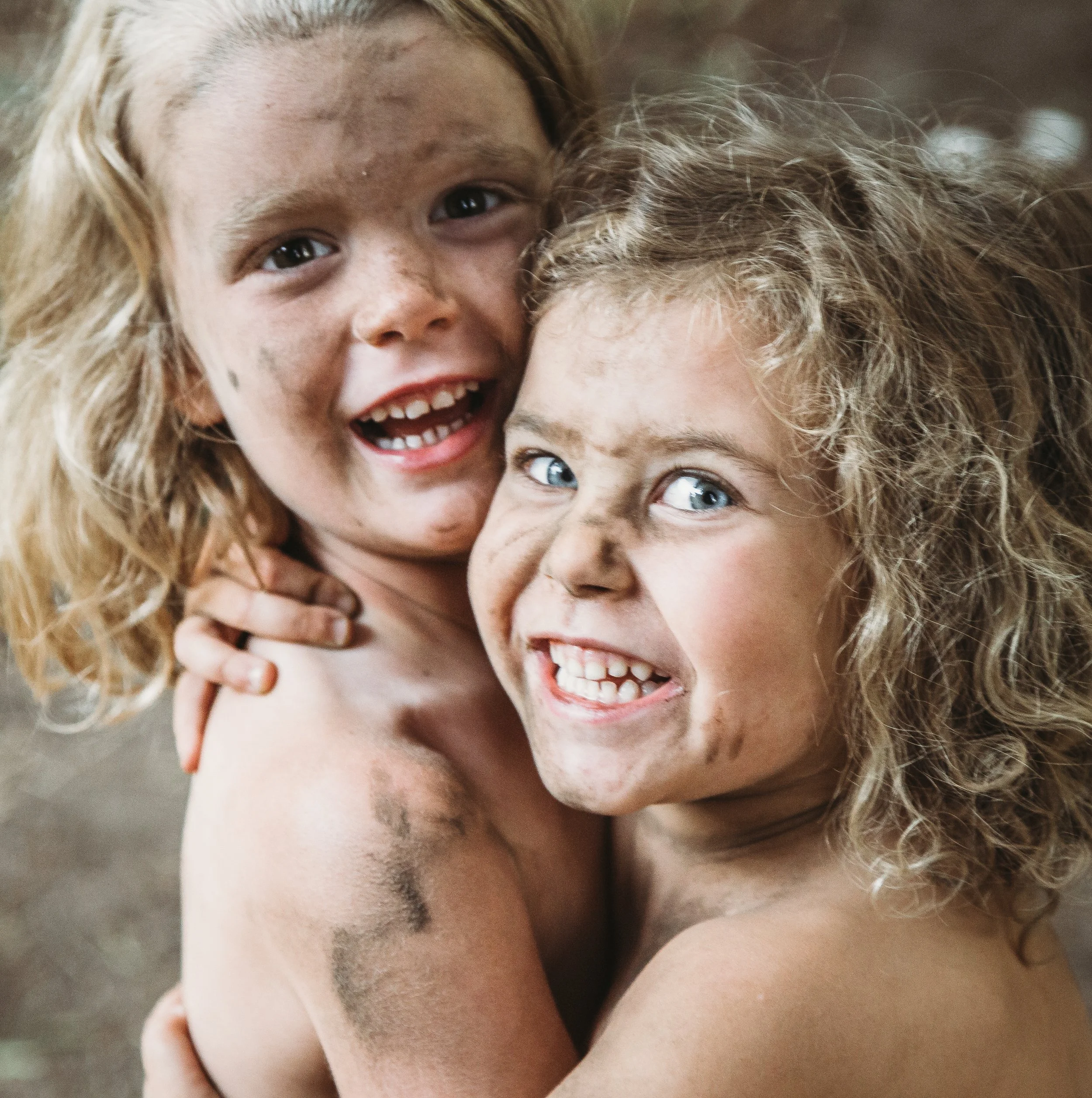 Two young children with curly hair and dirty faces, smiling and hugging enthusiastically.