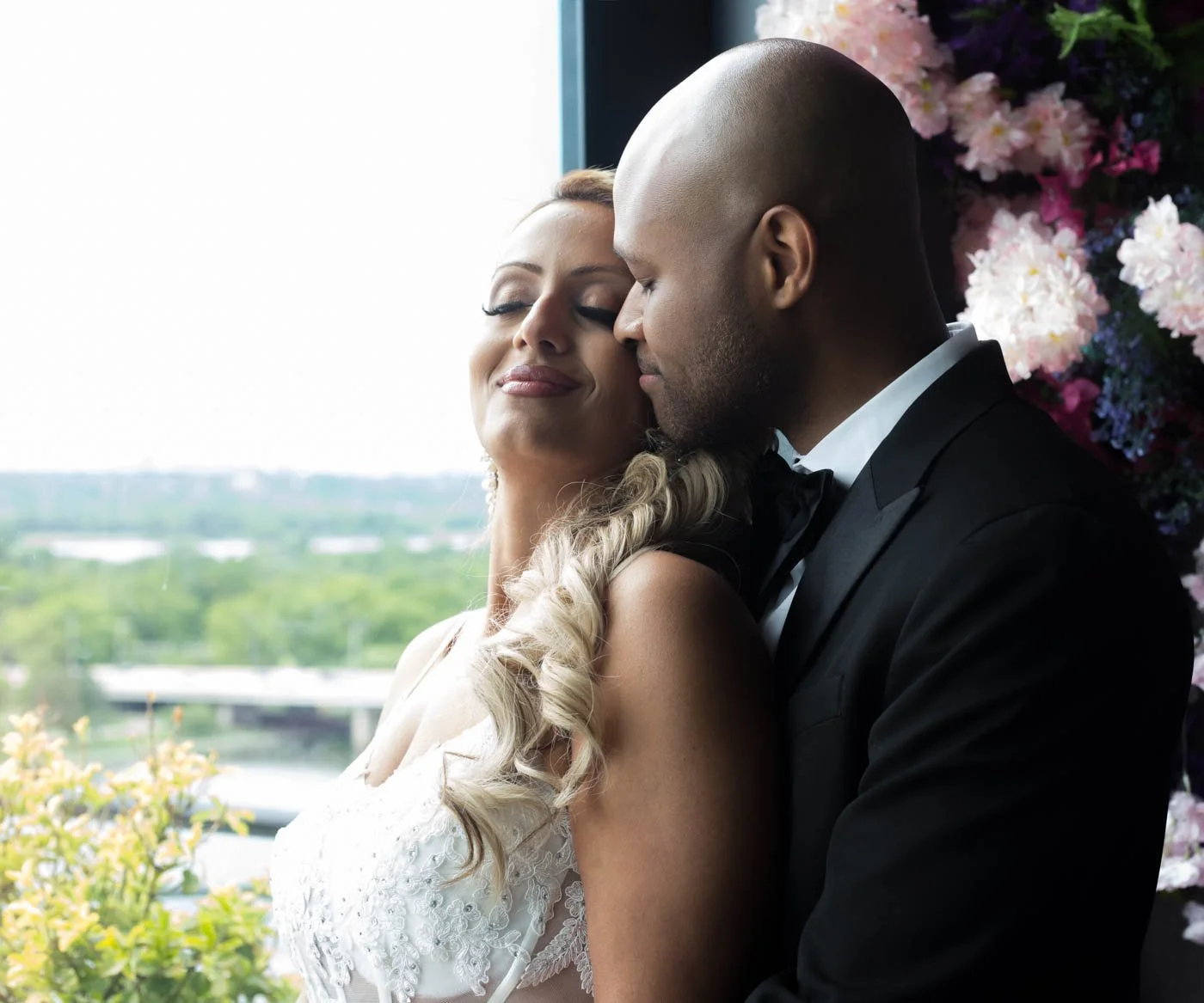 Bride and groom embracing near window with floral backdrop.