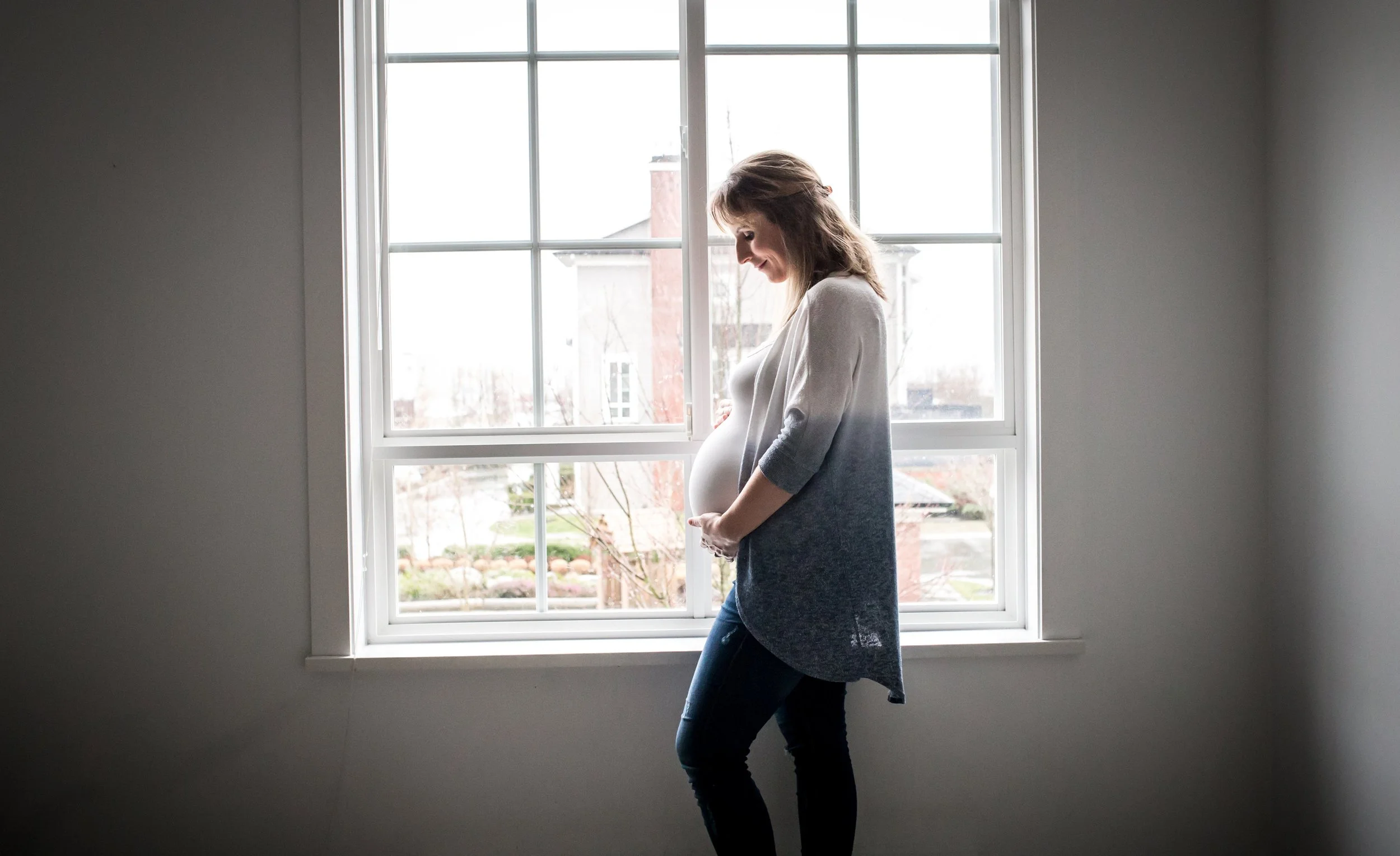 Pregnant woman smiling standing by a window, holding her belly, in a bright room.
