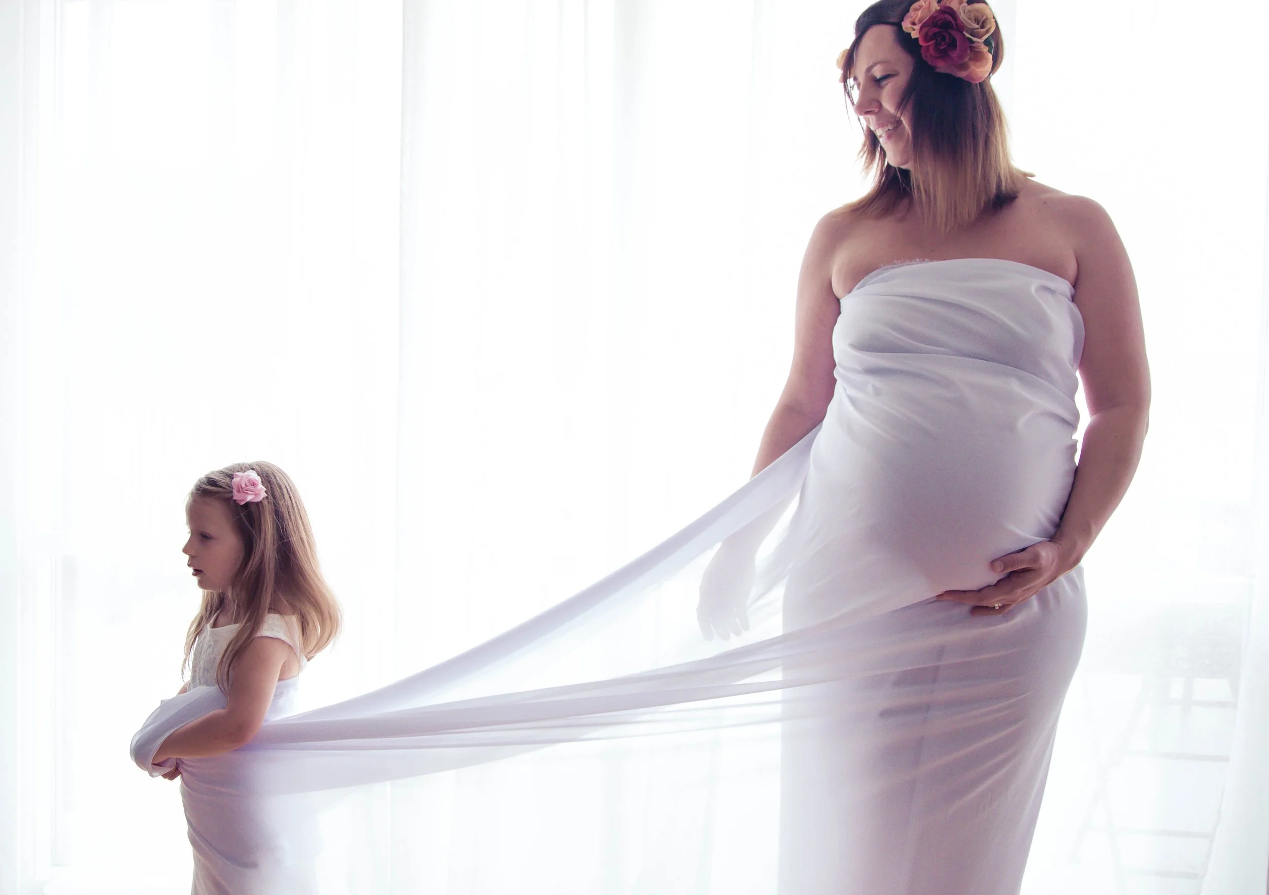 Pregnant woman and young girl in white dresses, holding sheer fabric, flower accessories.