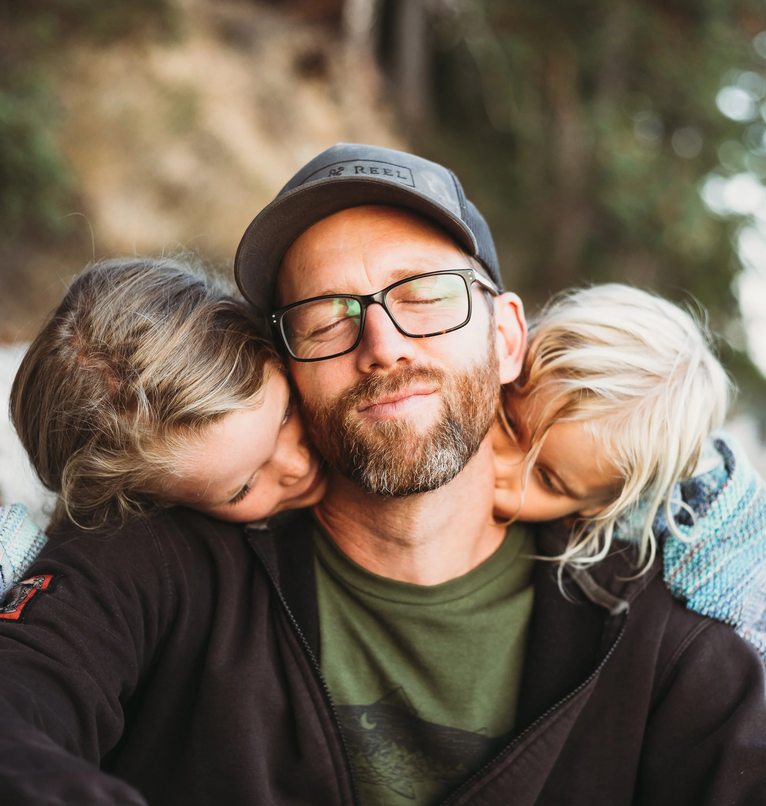 Man wearing glasses and a cap with two children hugging him affectionately against a blurred outdoor background.