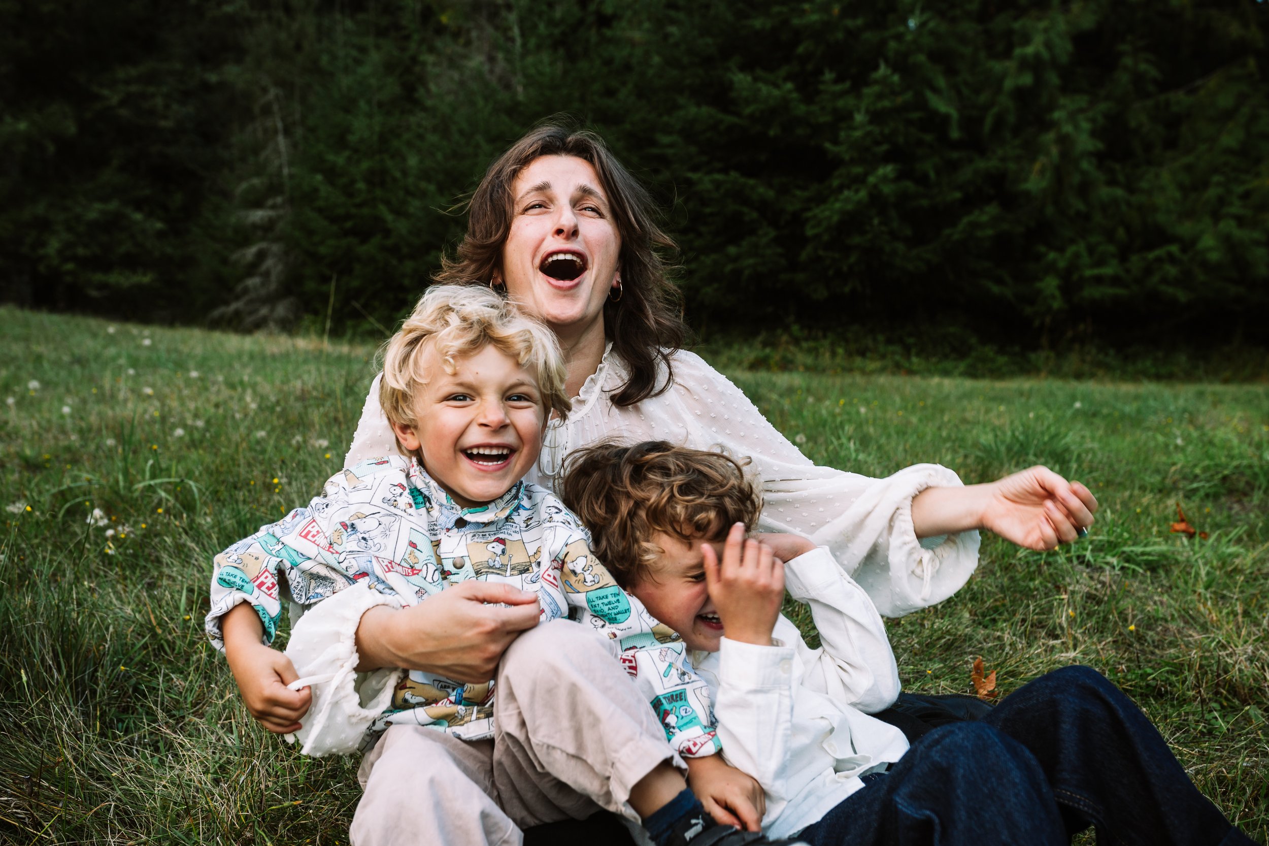 A woman and two children laughing while sitting on grass in a park.