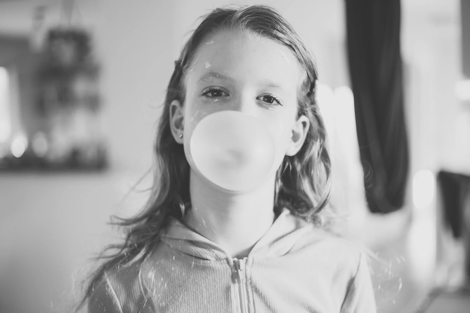 Black and white photo of a girl blowing a bubble with chewing gum, wearing a hoodie.