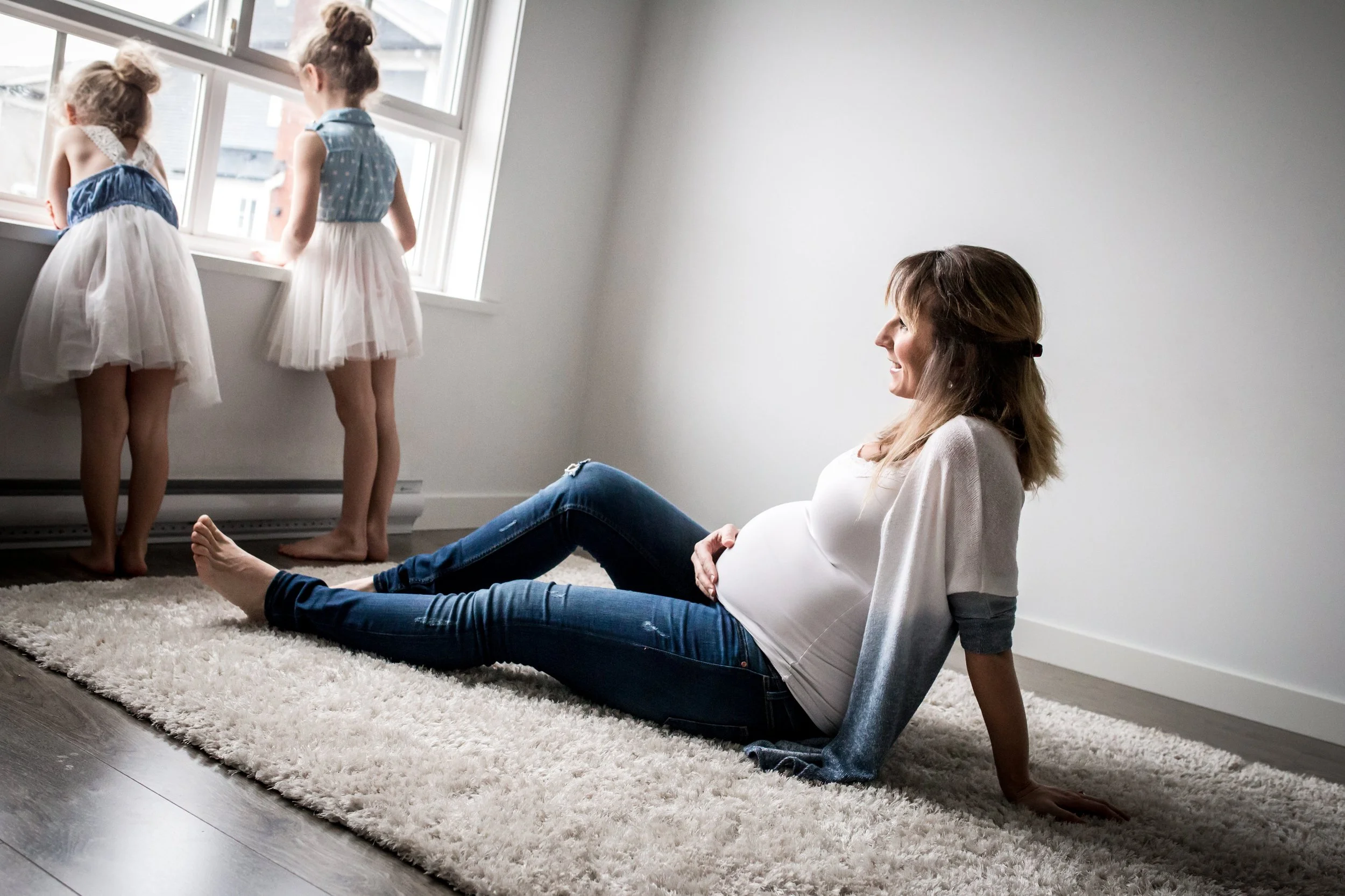 Pregnant woman sitting on carpet, two children in dresses looking out the window in a bright room.
