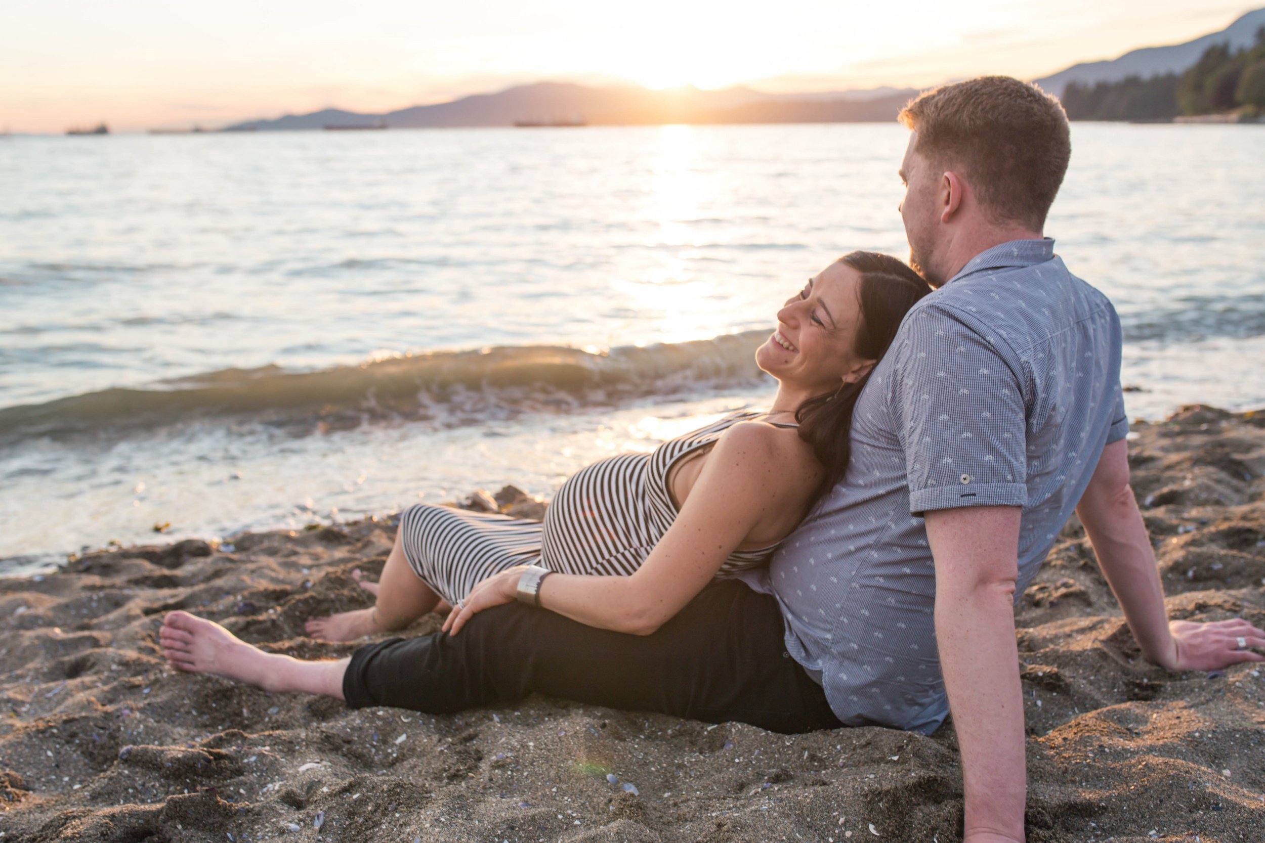 Pregnant woman in striped dress sitting on a beach with man at sunset.