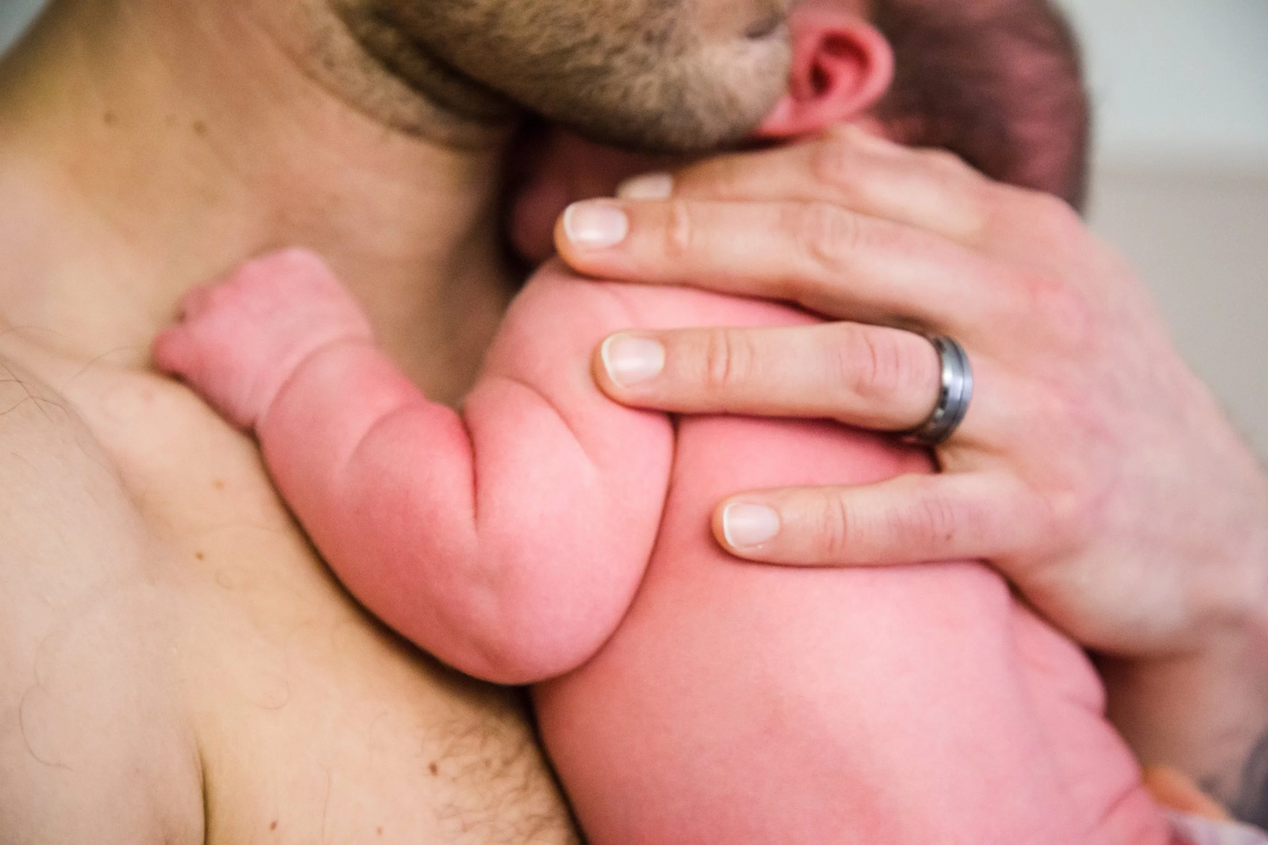 Close-up of a bearded man holding a newborn baby on his shoulder, with his hand gently resting on the baby's back.