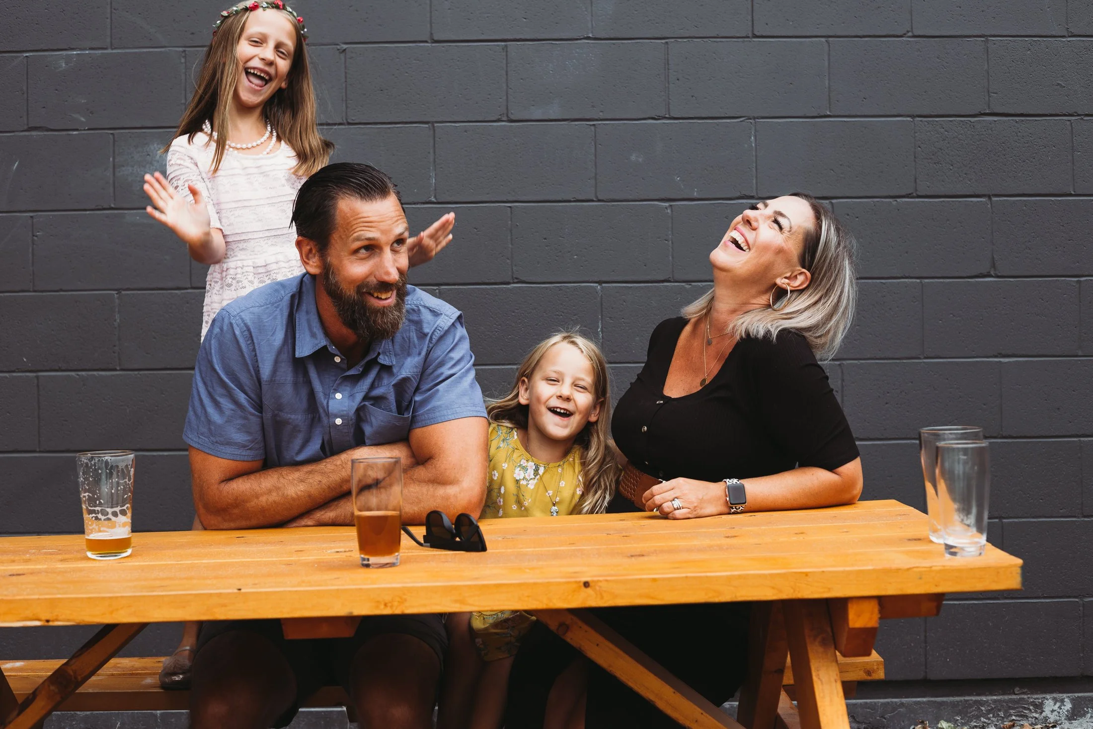 Happy family of four sitting at a wooden table outdoors, laughing and smiling in front of a brick wall.