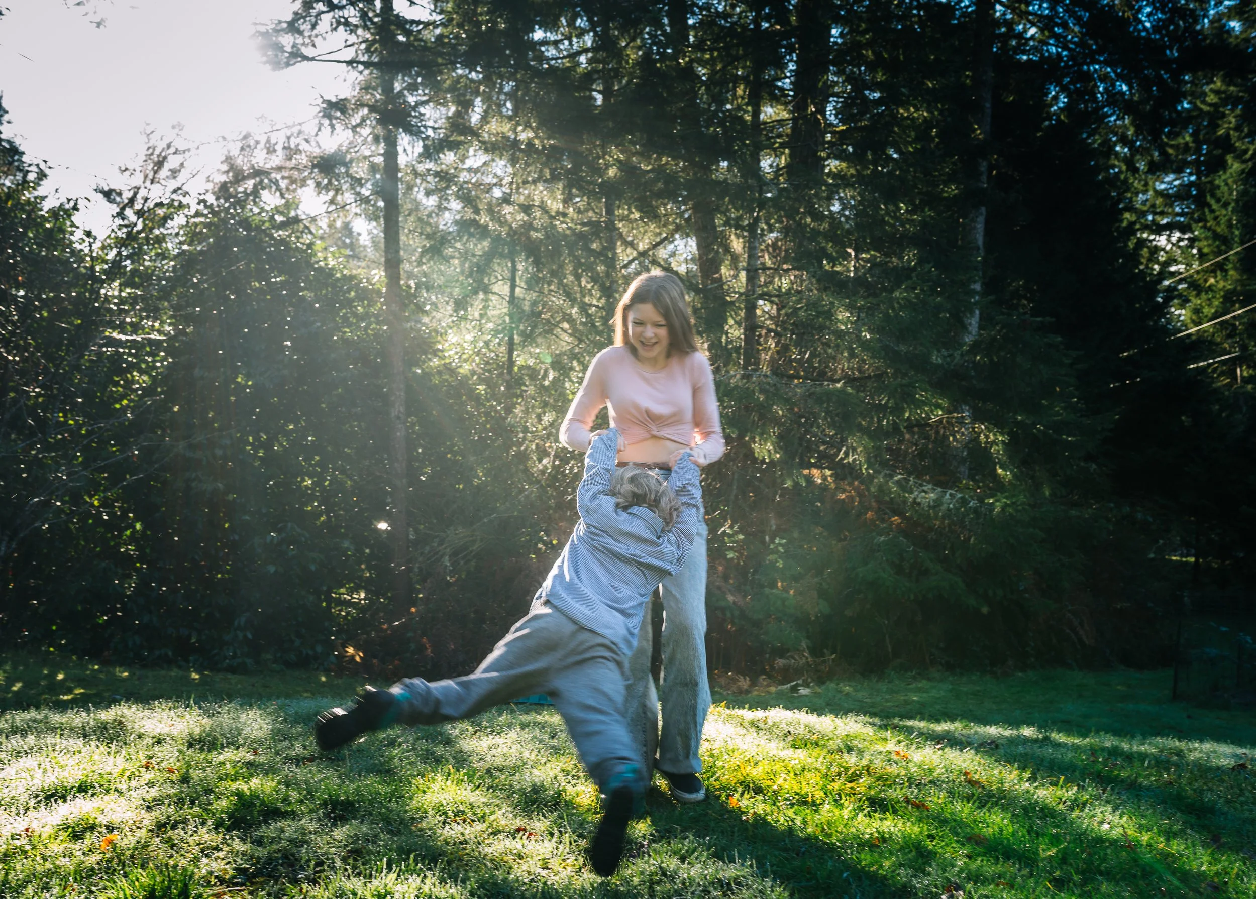Person spinning a child in a sunlit grassy area with trees in the background.