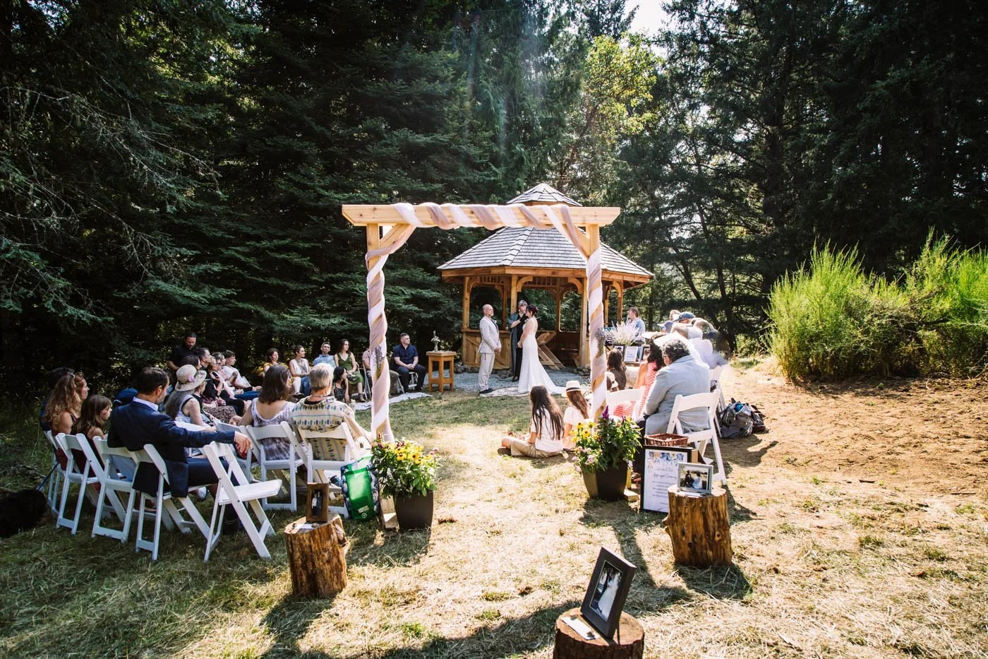 Outdoor wedding ceremony in forest setting with wooden arch and guests seated on white chairs.