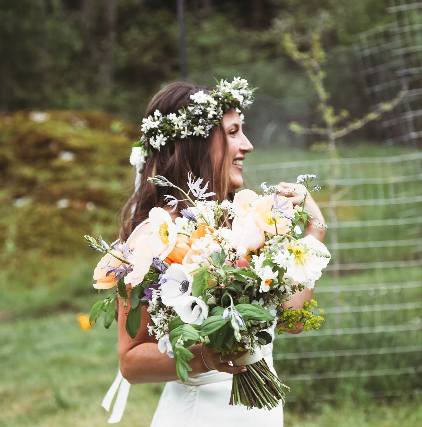 Woman in white dress holding a colorful bouquet of flowers, wearing a floral crown, smiling, outdoors.