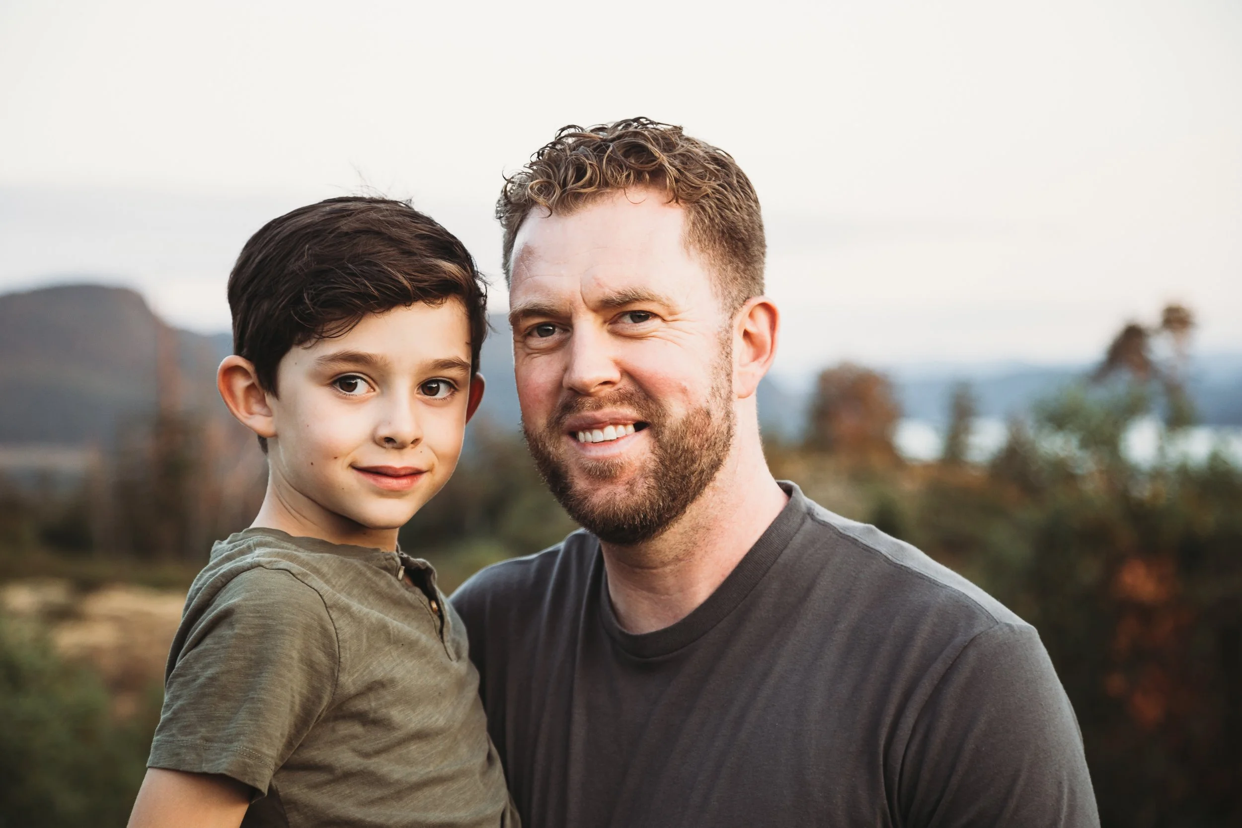 A man and a boy standing together outdoors, smiling, with a scenic background of mountains and trees.