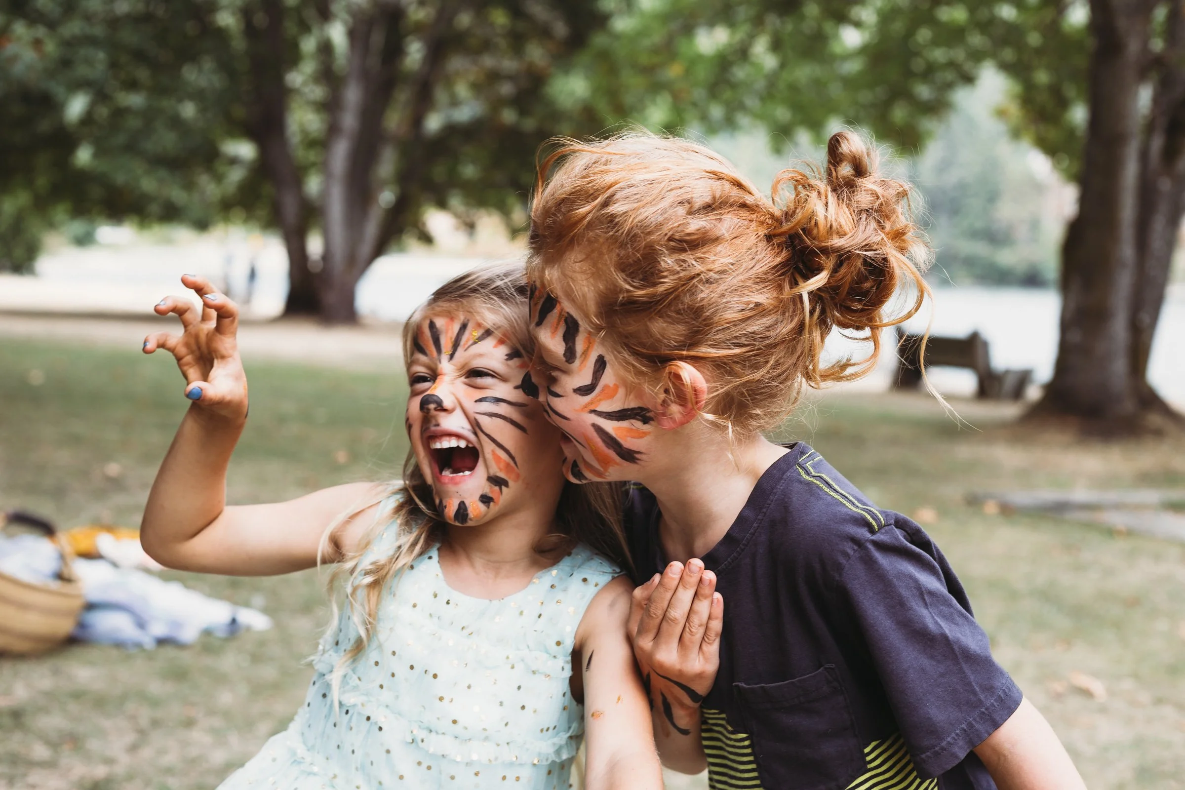 Two children with face paint designed to look like tigers, embracing and playing in a park with trees in the background.