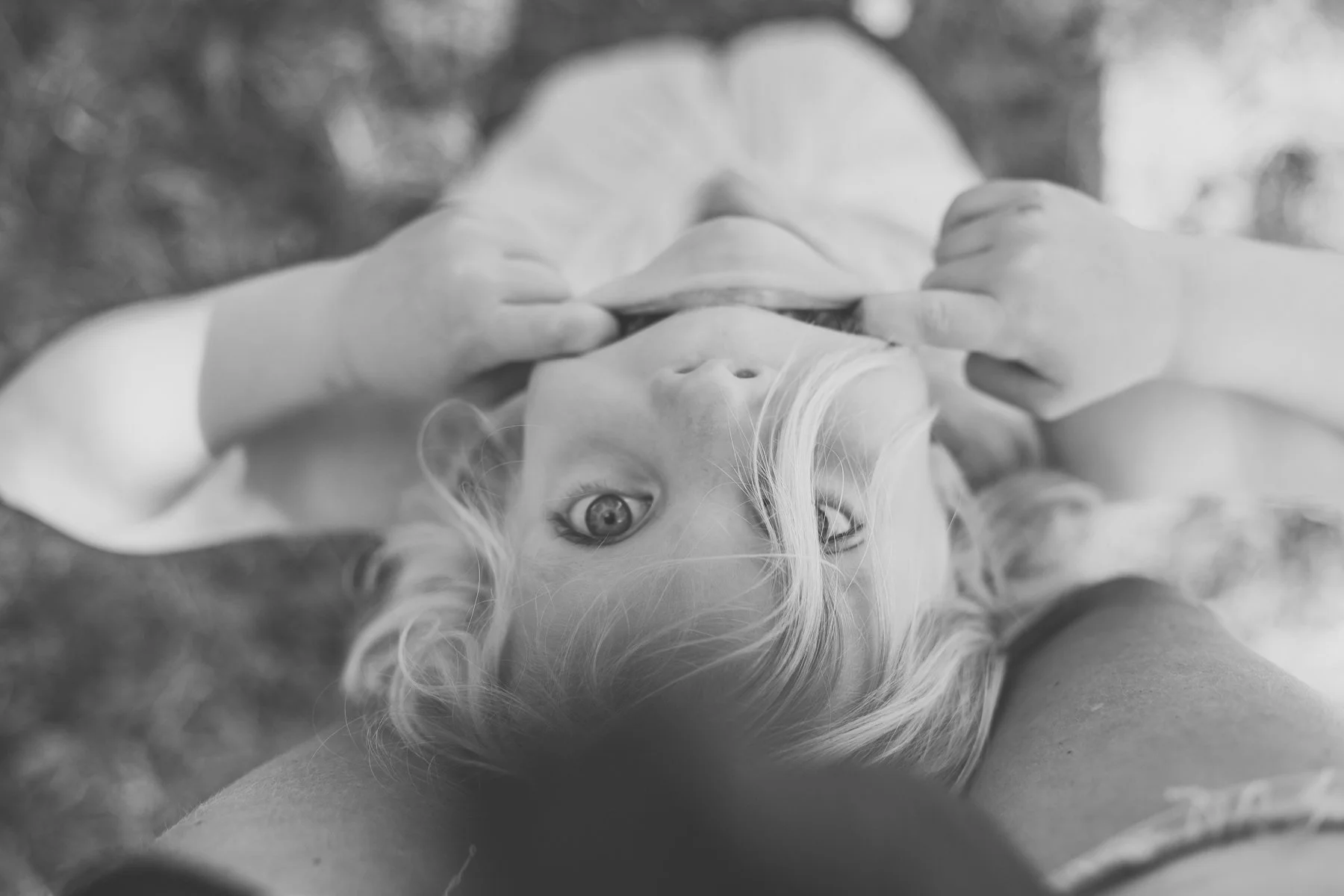 Black and white photo of a child with blonde hair peering up, playfully pulling their mouth with fingers.