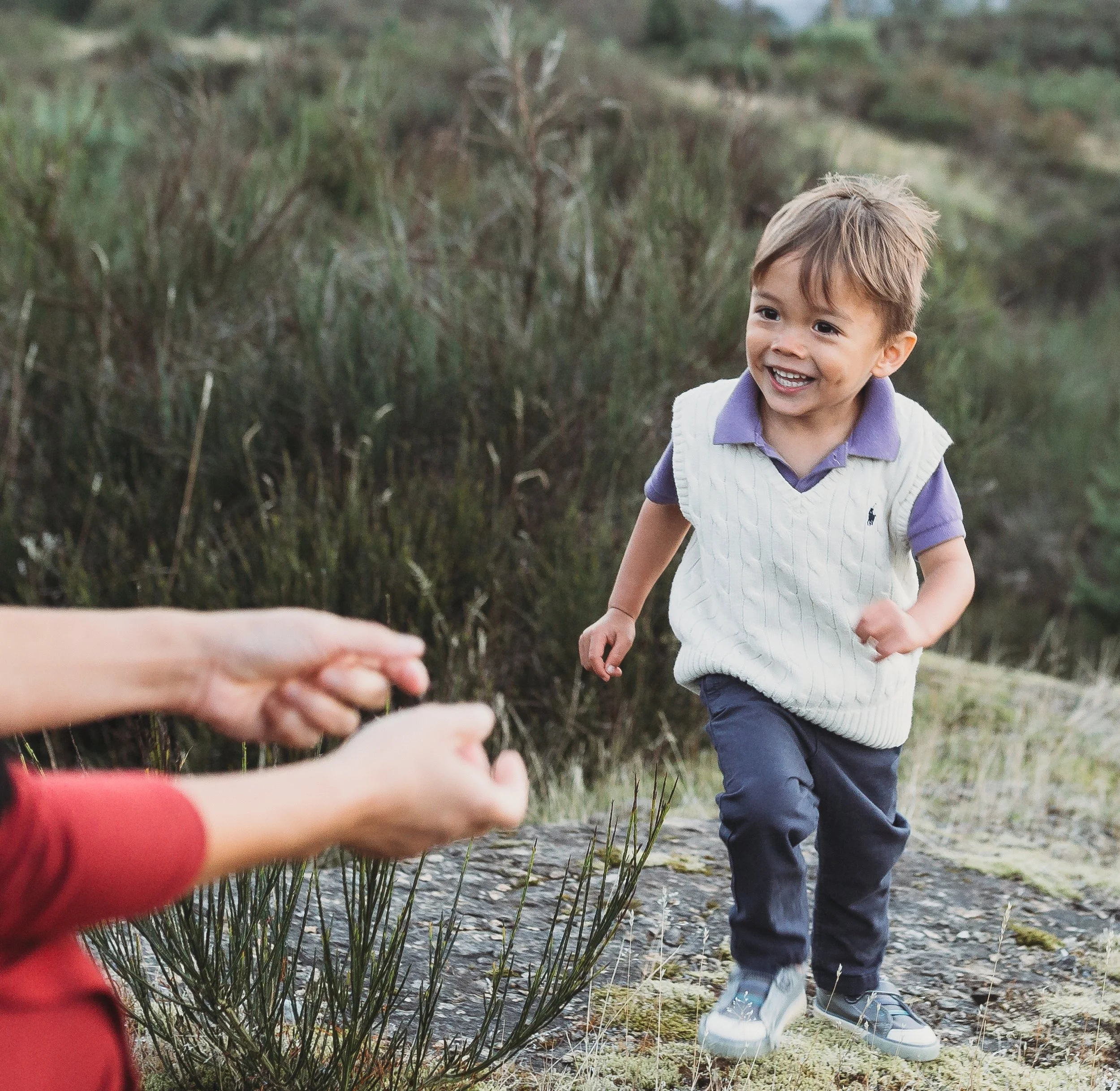 Smiling toddler in a white sweater vest and purple shirt running outdoors near a person extending their hands.