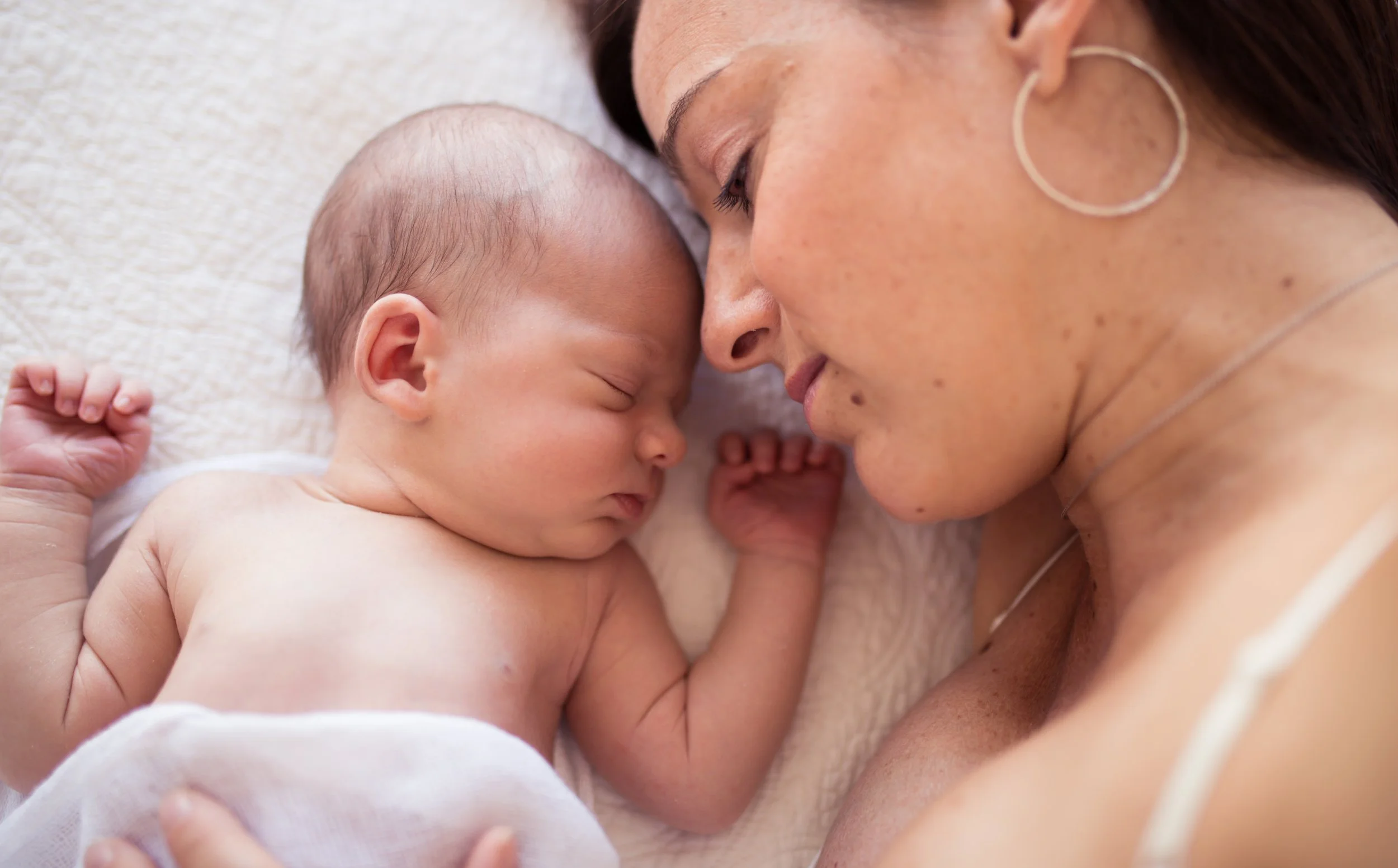 Mother and newborn baby lying close together on a white blanket, both sleeping peacefully.