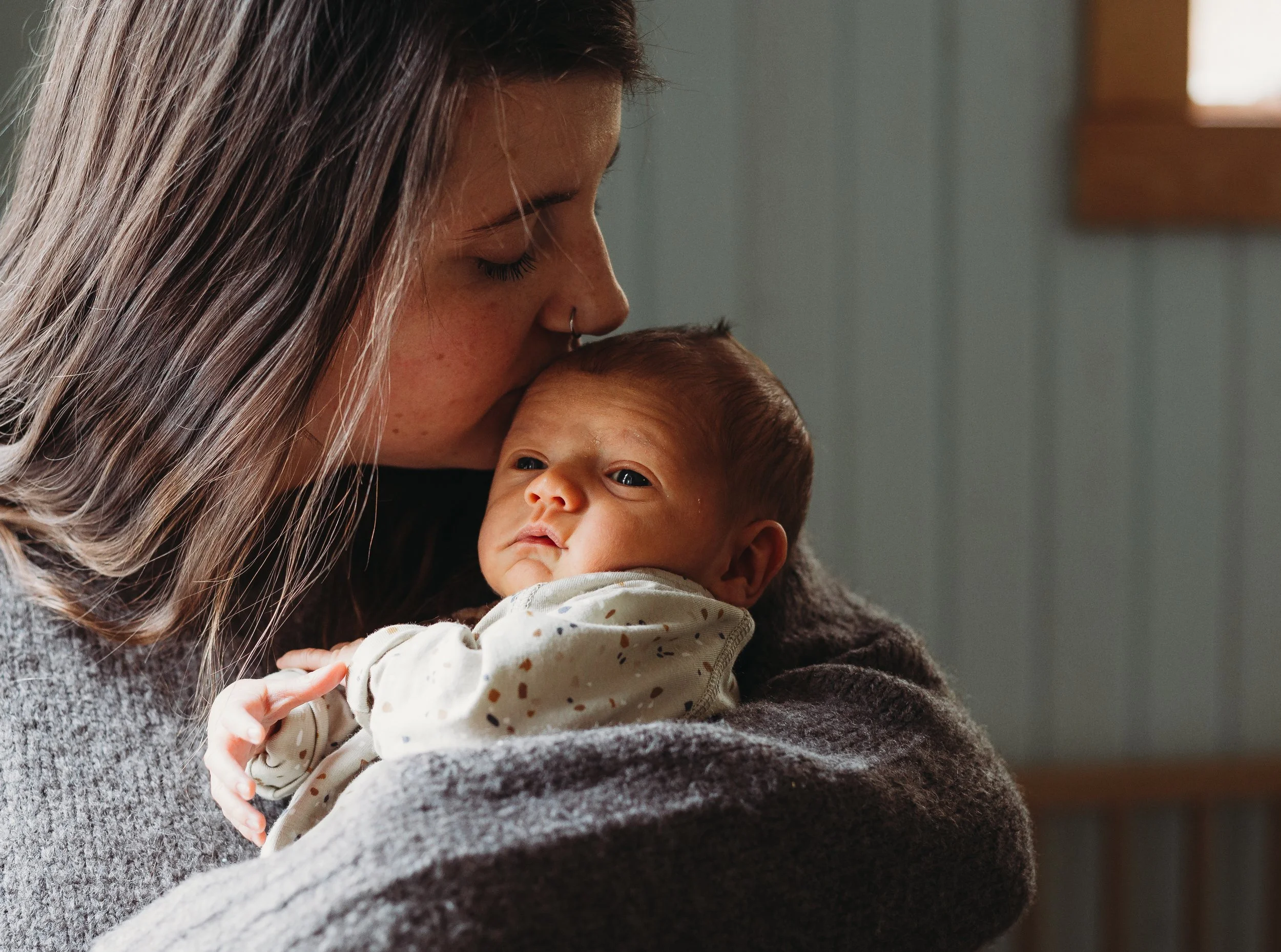 Mother kissing infant's head indoors, baby wrapped in blankets.
