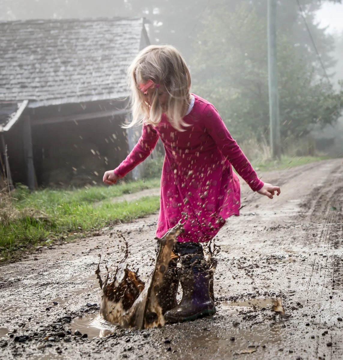 Child splashing in a muddy puddle on a dirt road, wearing a pink dress and boots.