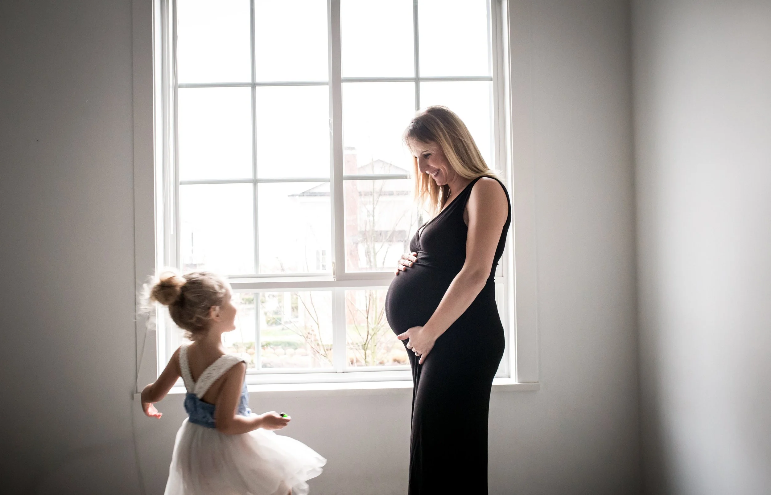 Pregnant woman in black dress smiling at child in dress by window