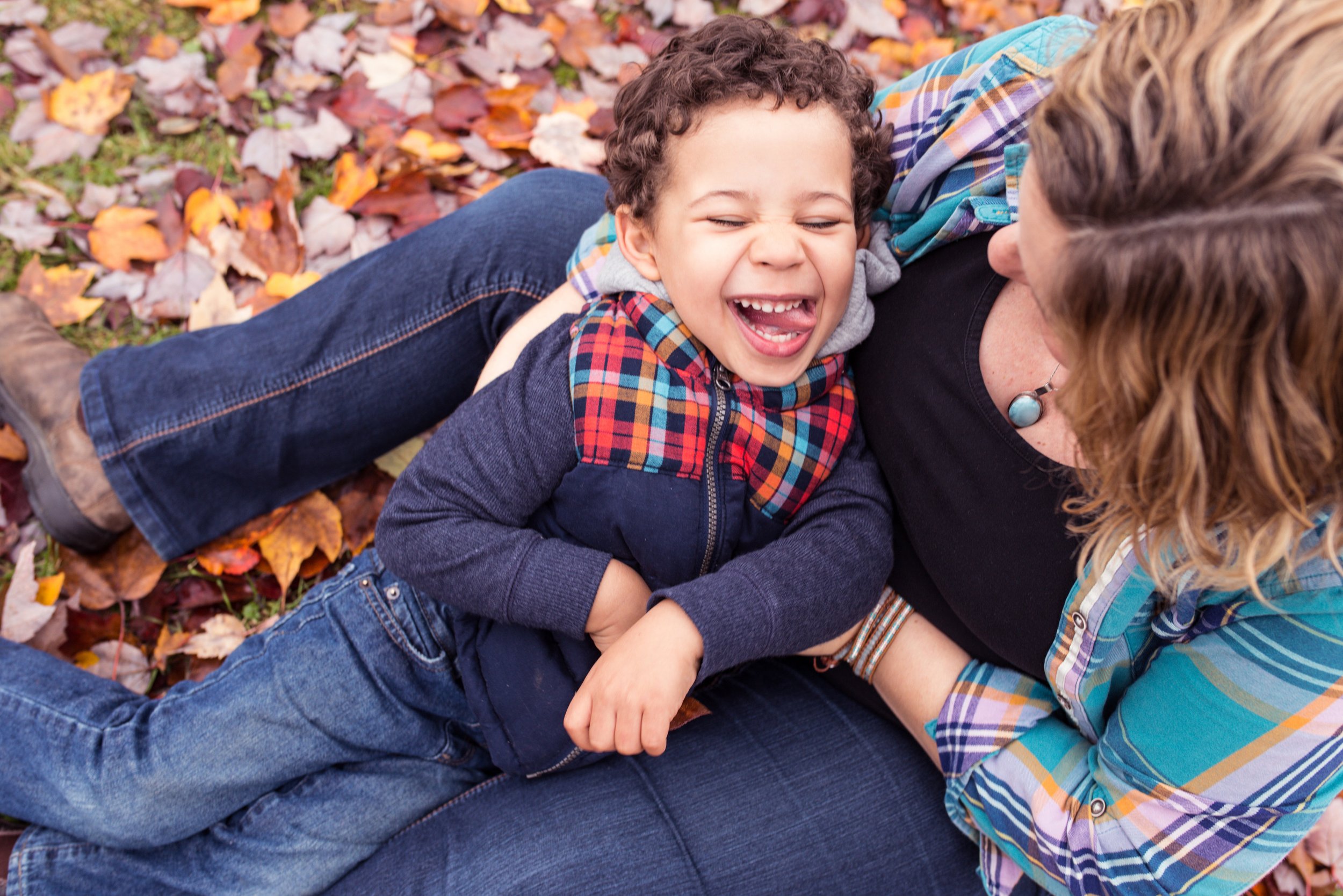 A mother and child sitting on autumn leaves, the child laughing joyfully while being hugged by the mother.