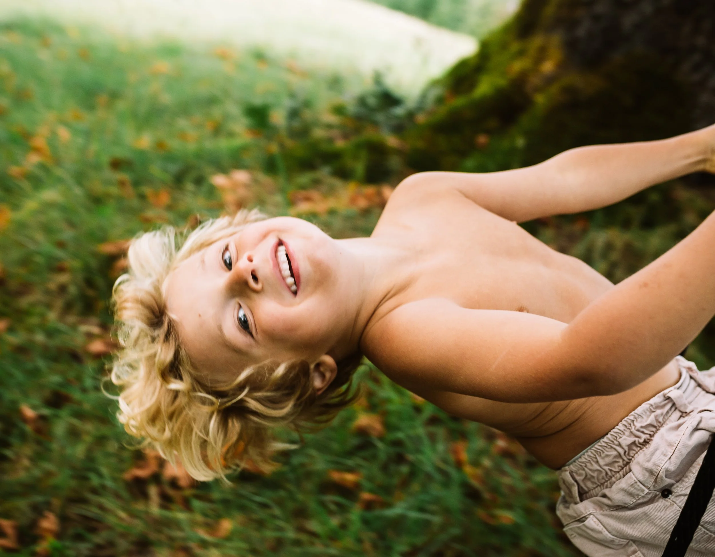 A child with blonde hair swinging outdoors on a rope swing, surrounded by greenery.