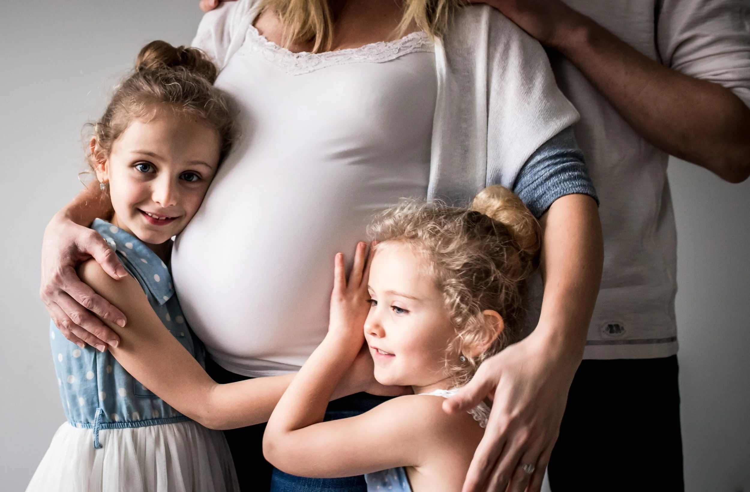 Two young girls hugging a pregnant woman's belly, surrounded by another adult's hands, symbolizing family and maternal love.