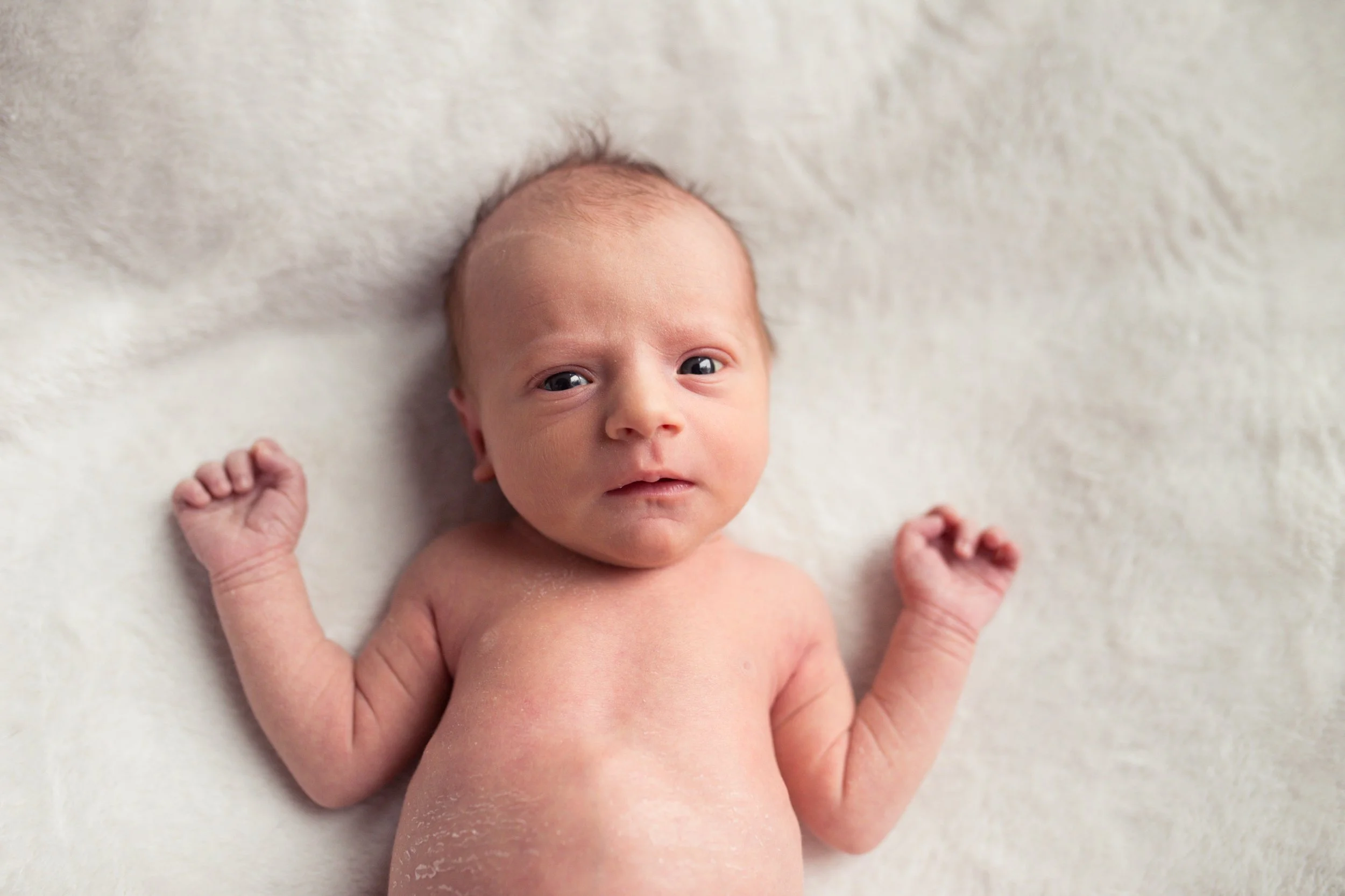 Newborn baby lying on a soft white blanket, looking at the camera with arms slightly raised.