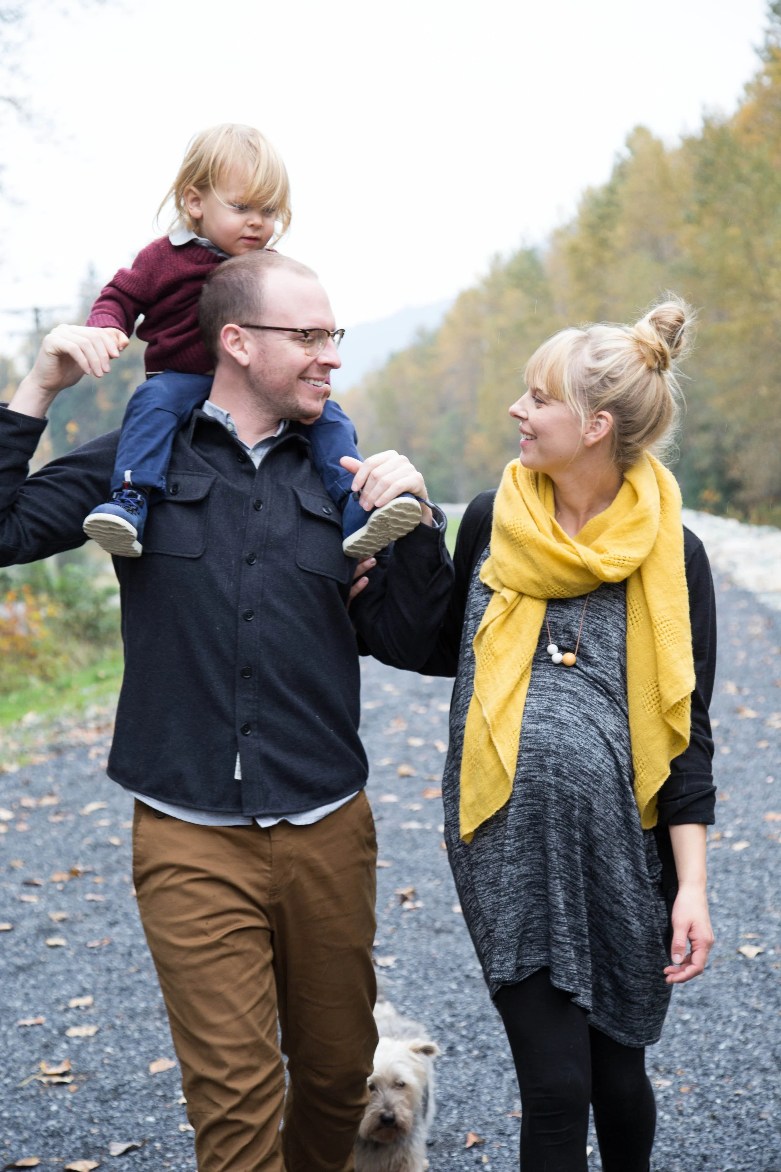 Family walking outdoors; man carrying child on shoulders, woman wearing yellow scarf, dog nearby.
