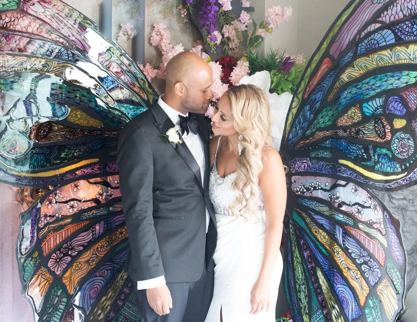 Couple in formal attire standing in front of colorful butterfly wings backdrop.