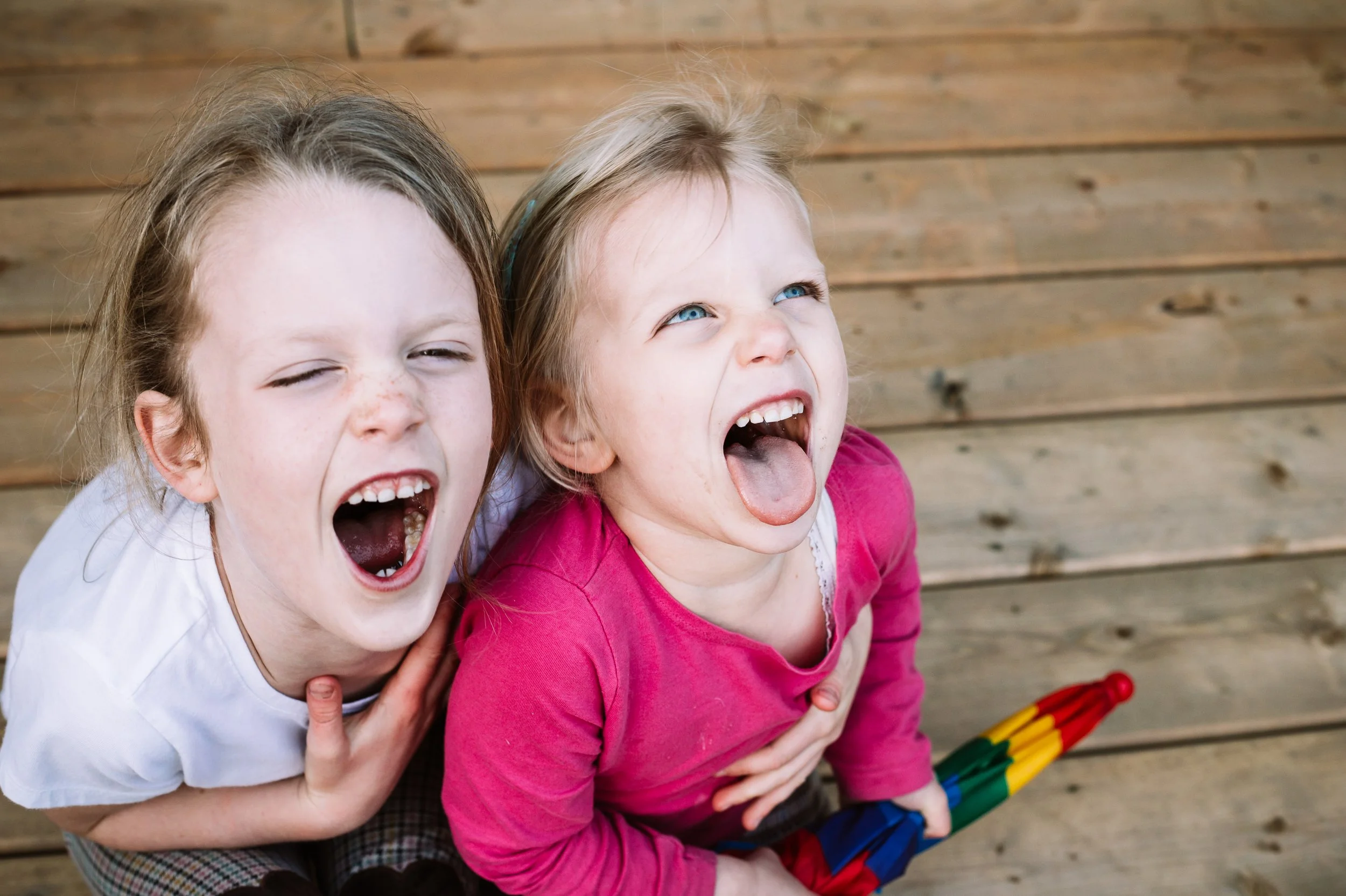 Two children laughing with mouths open, one holding a colorful umbrella on a wooden floor.
