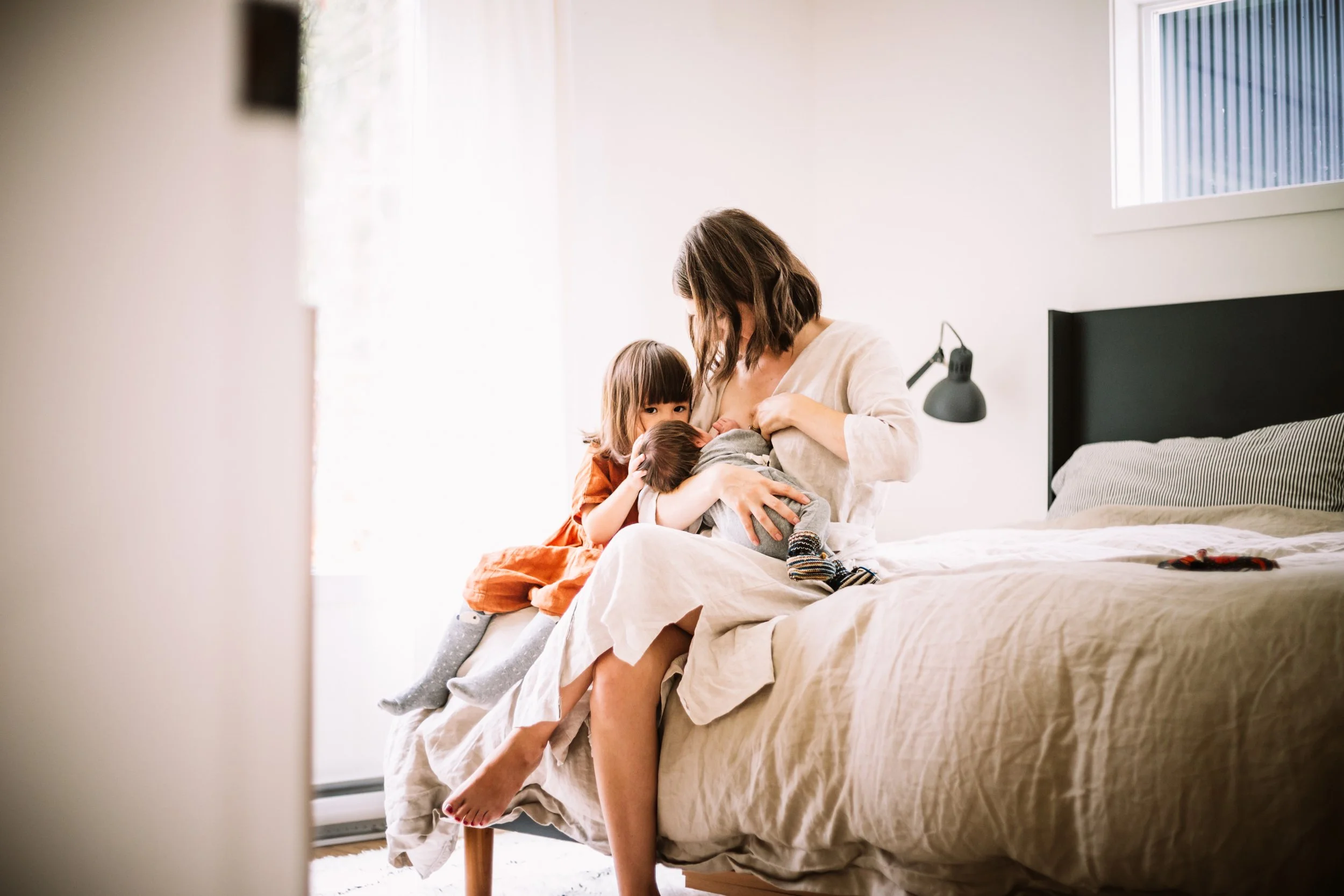 Mother breastfeeding baby with child sitting beside on bed in cozy bedroom.