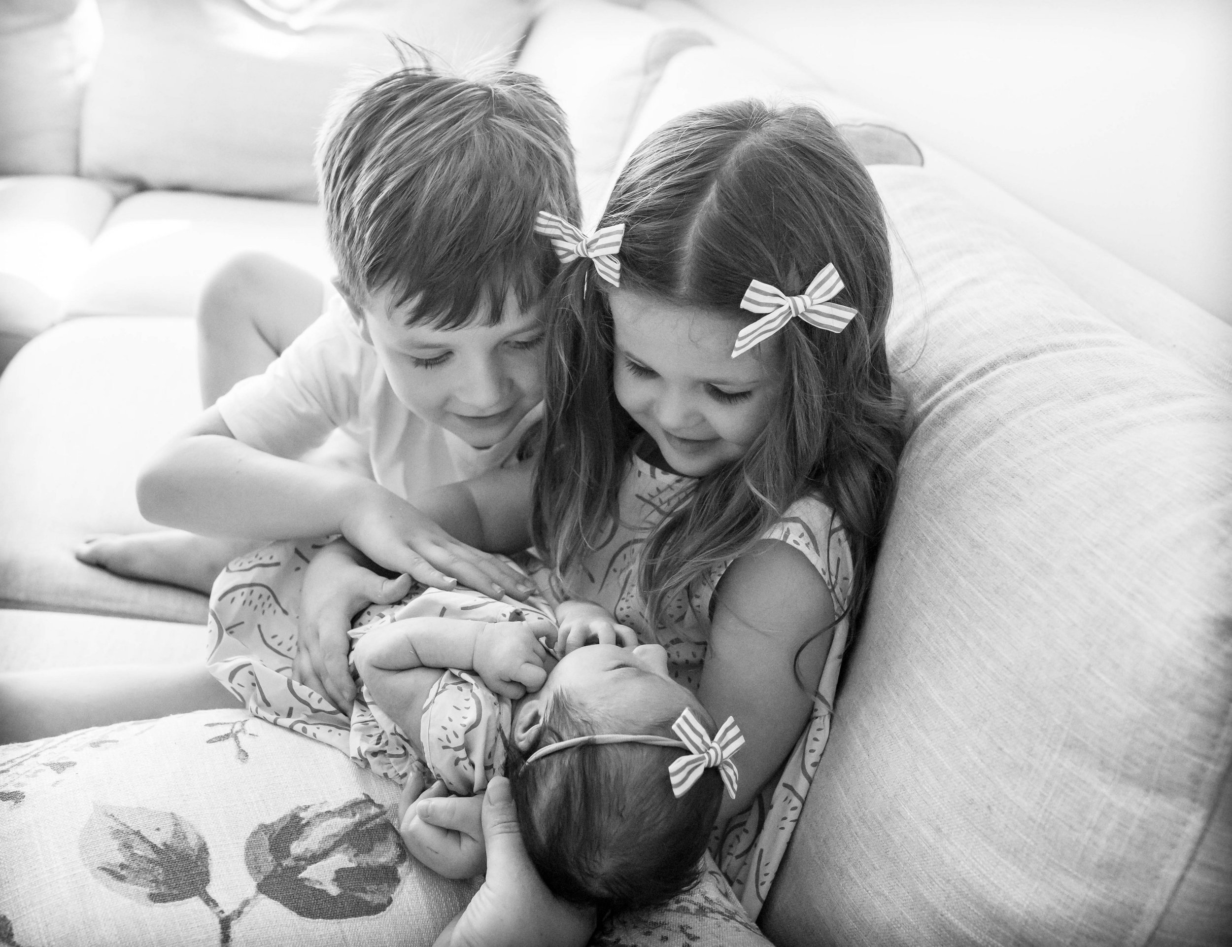 Black and white photo of two young children with a baby on a sofa, the girl holding the baby, both children wearing matching hair bows.
